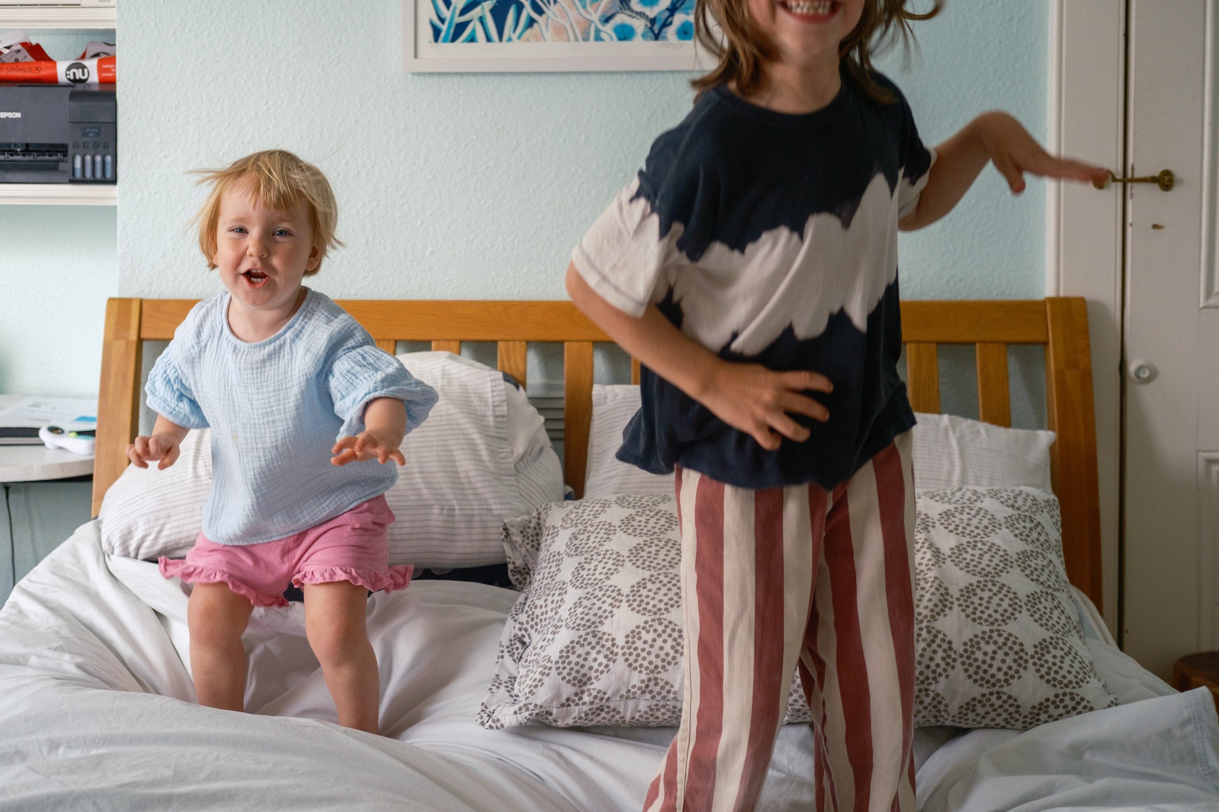 Two children are on a bed, one is standing and the other is jumping. The child on the left wears a light blue top and pink shorts, with a joyful expression. The child on the right, wearing a navy and white tie-dye shirt and striped pants, is partiall