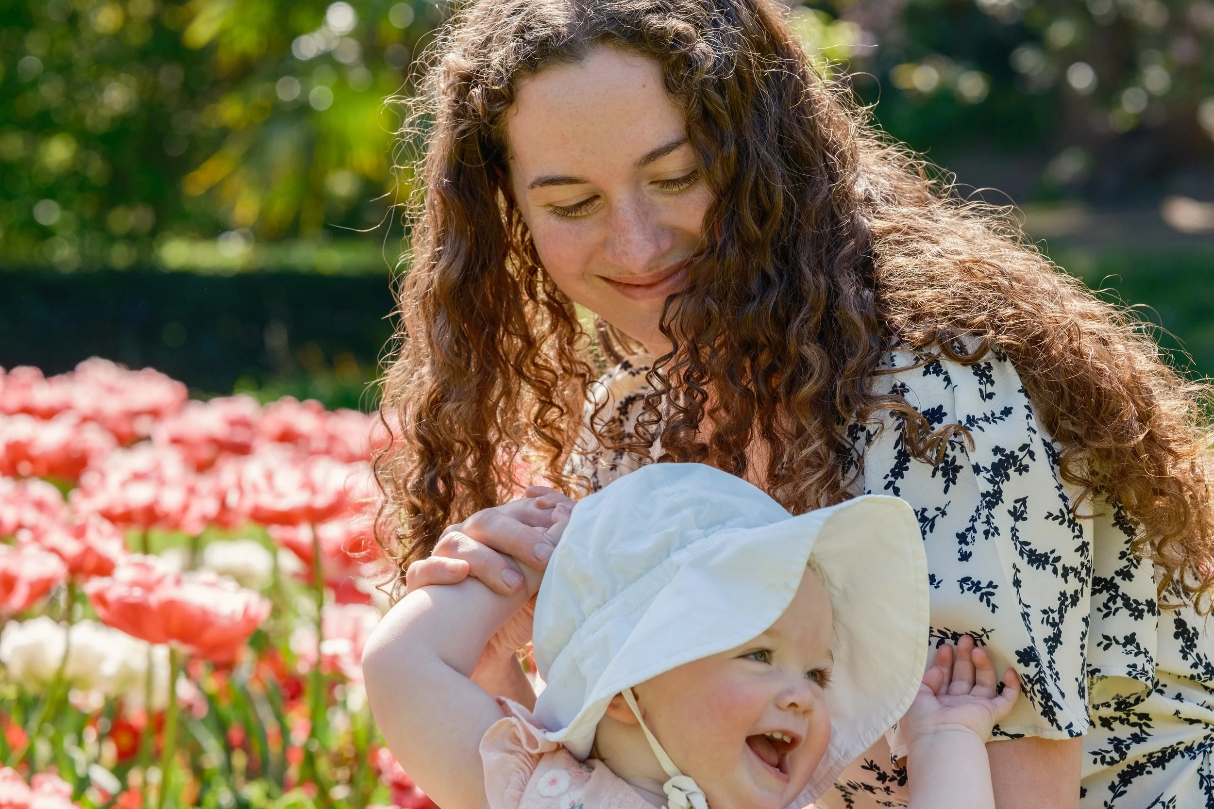 A woman with curly hair and two young children, one with a sun hat the other with a pink outfit, outdoors among colorful flowers, smiling and enjoying a sunny day.
