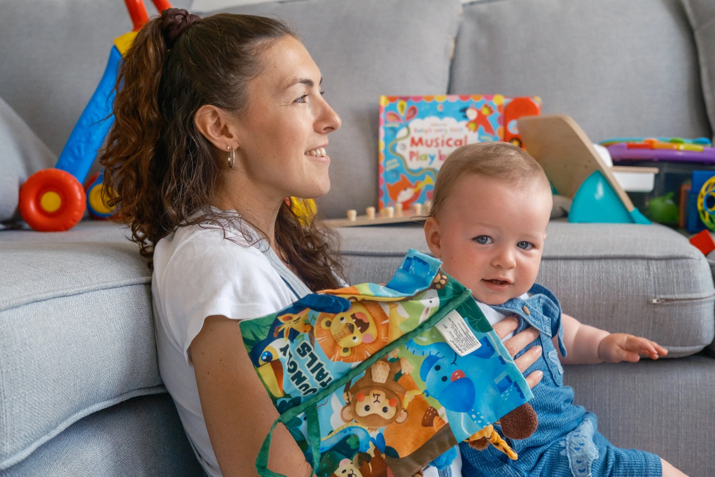 A woman with curly brown hair, wearing a white shirt, is sitting on a gray couch holding a young boy with short light brown hair, blue eyes, wearing a denim shirt and shorts, and a colorful cloth bag with animal designs. Behind them, toys and a child