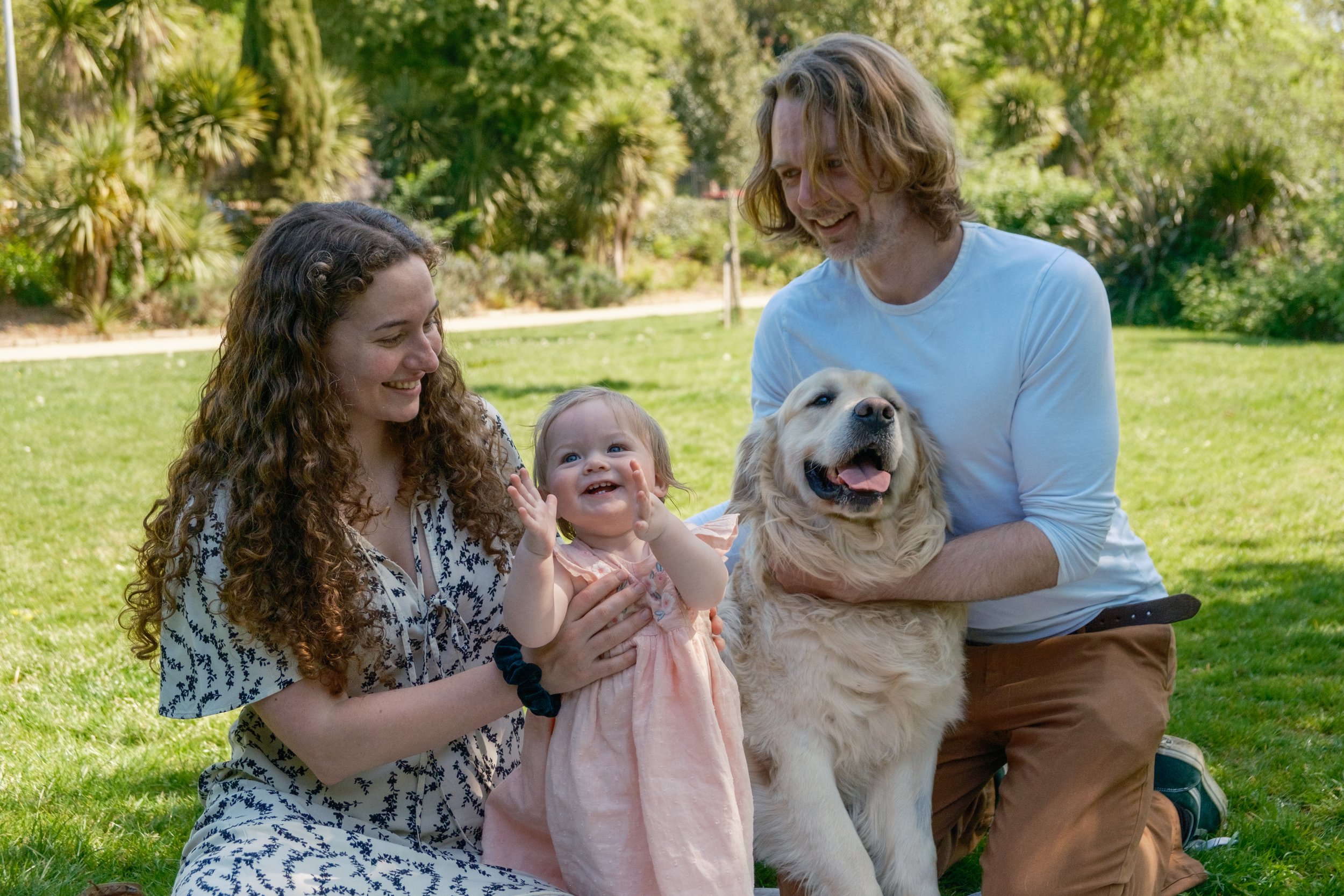 A family enjoying a day outdoors with a dog and a small child. The woman with curly hair holds the small child who is smiling and clapping. The man with shoulder-length hair holds a happy golden retriever. They are on a grassy area with trees in the 