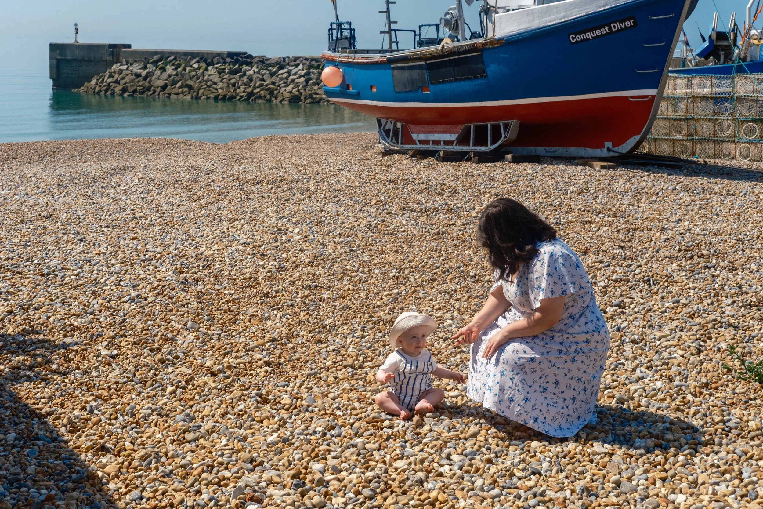 A woman and a young child sitting on a pebble beach near a boat and a breakwater, with the woman reaching out to the child, who is wearing a hat and striped outfit.