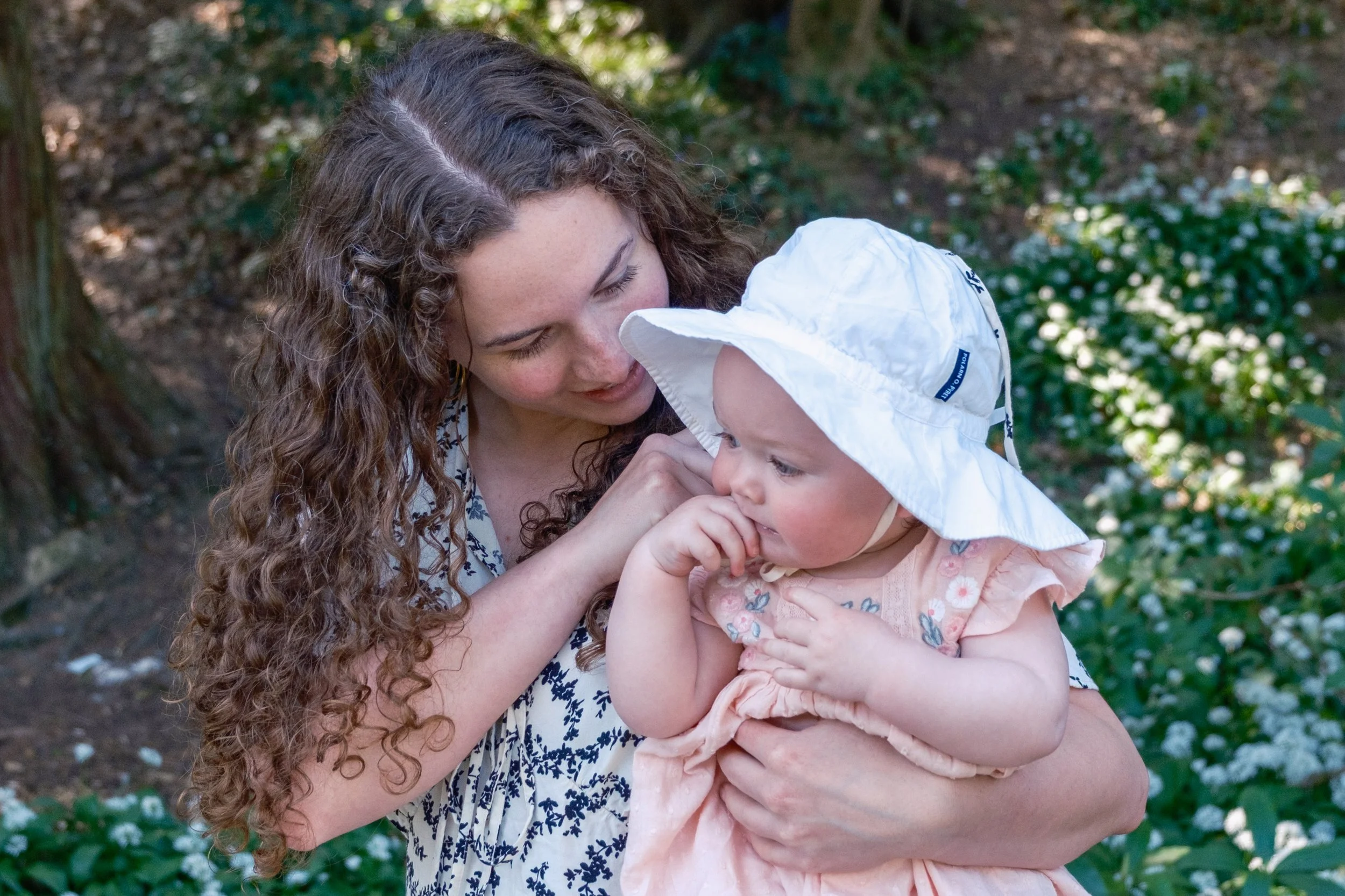 A woman with curly brown hair holding a young girl wearing a white sun hat outdoors surrounded by green bushes and white flowers.