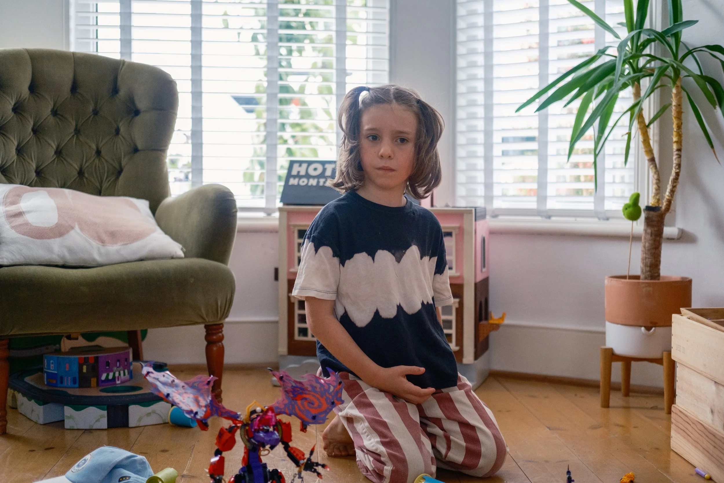 A young girl with brown hair in pigtails sits on her knees on a hardwood floor in a room with large windows and blinds. She is wearing a navy and white tie-dye shirt and red and white striped pajama pants. Toys and a red and purple robot figurine are