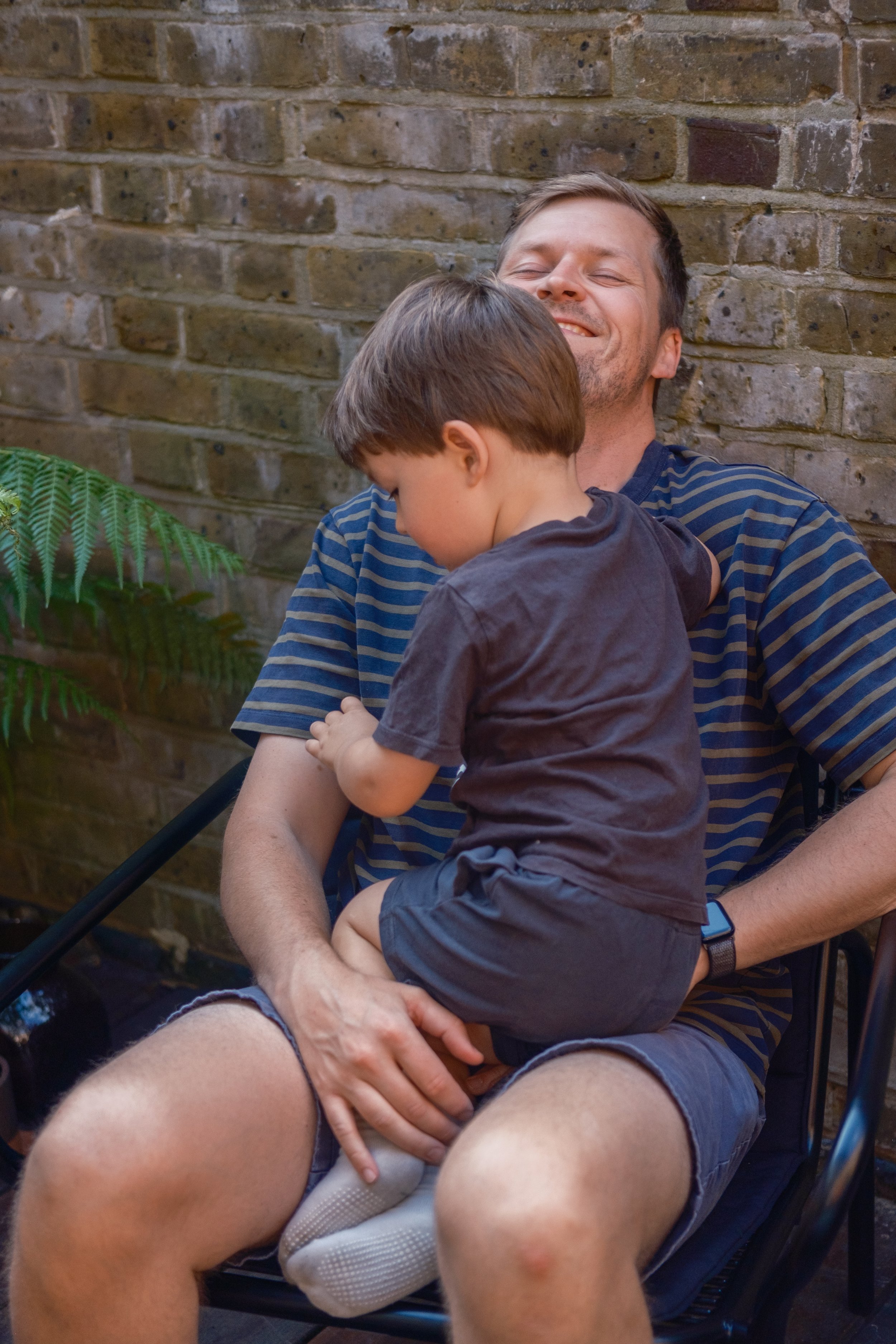 A man sitting outdoors against a brick wall, smiling with his eyes closed, holding a young boy on his lap. The boy is leaning forward with his head resting on the man's chest.
