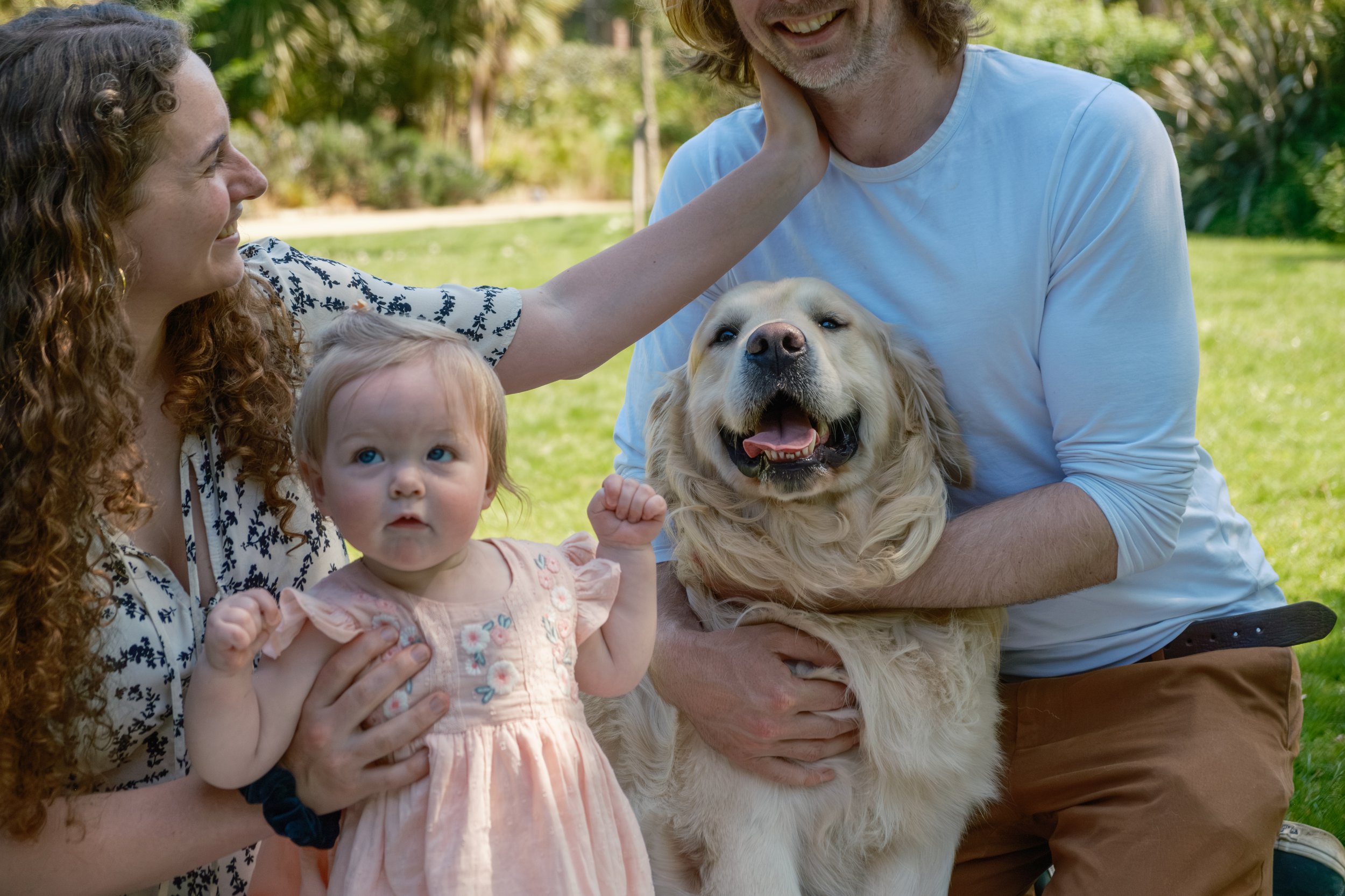 A family of two adults, a young girl, and a dog outdoors in a park. The woman has curly hair and is smiling at the dog, while the man is holding the dog. The young girl has light hair and is looking at the camera with raised fists. The dog, a golden 