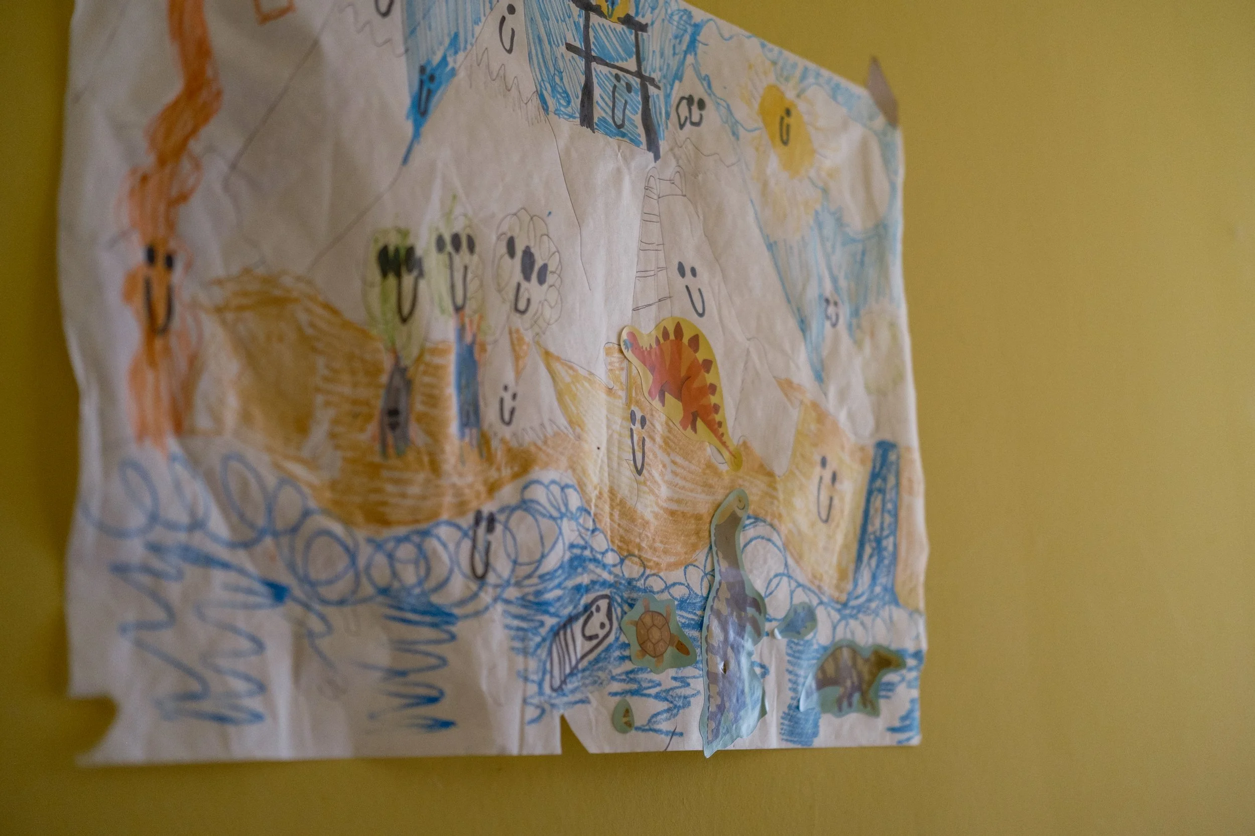 Colorful children's drawing of a prehistoric landscape with dinosaurs and smiling characters, on paper attached to a yellow wall.