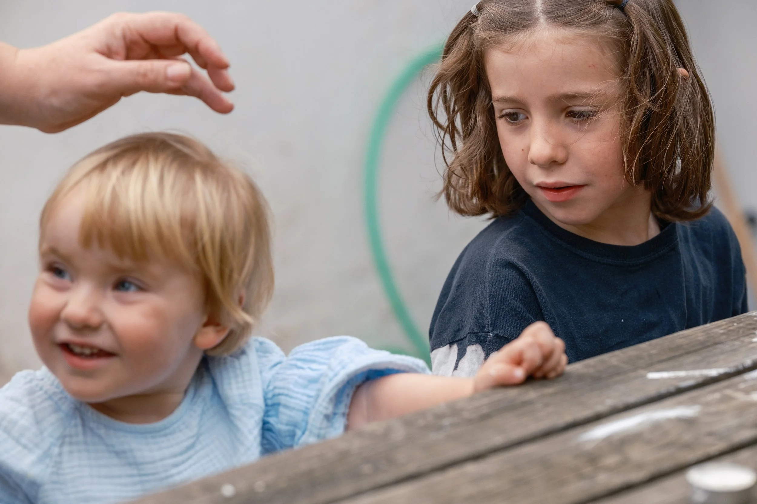 Two children, a young girl with curly brown hair and an older boy with blonde hair, are engaged in an activity at a wooden table. An adult's hand is reaching towards them, possibly to assist or guide. The children are smiling and seem to be focused o