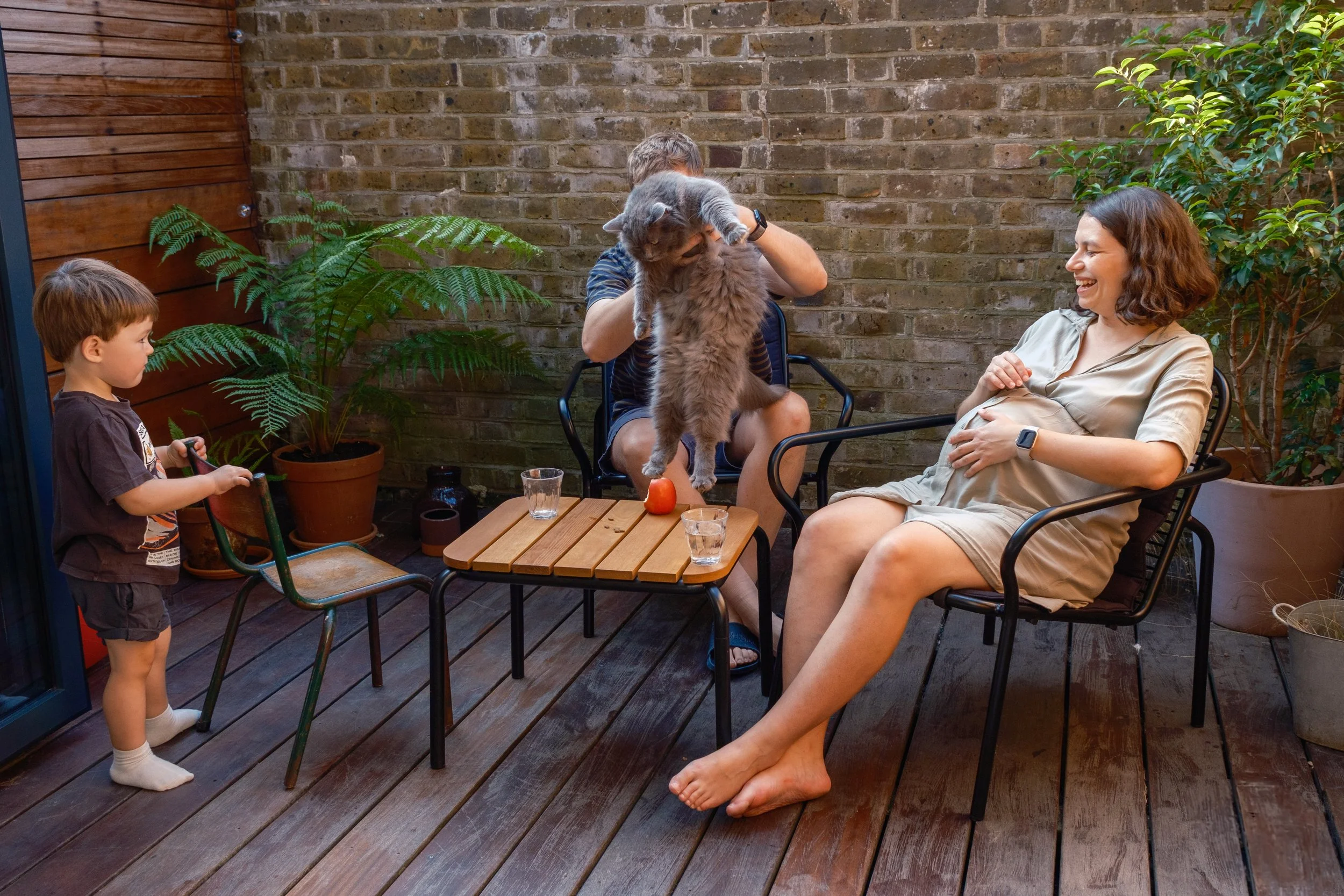 A family enjoying time in a backyard patio, with a woman smiling while sitting in a chair, a man holding a large gray cat over a small table, a young boy holding a shoe and a toy, and lush green plants against a brick wall.