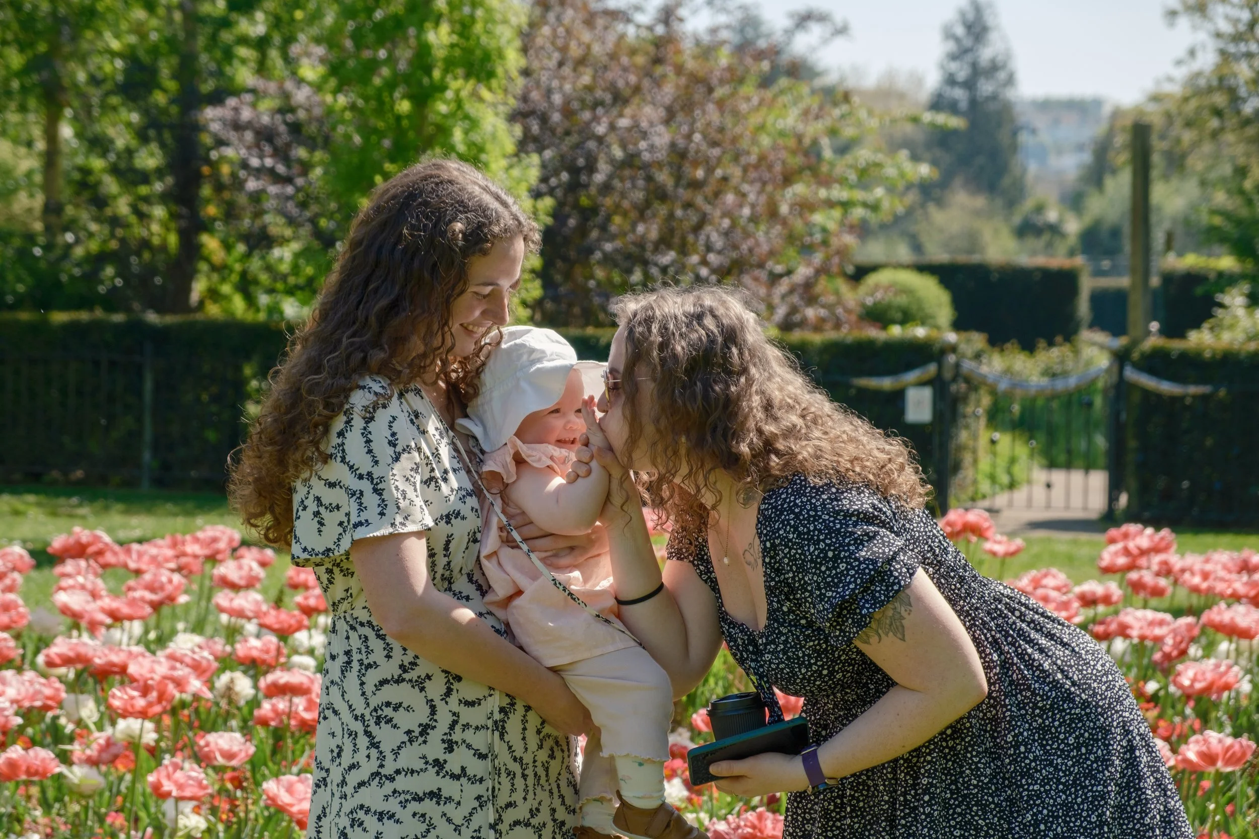 Two women and a baby in a garden with pink flowers, trees, and a gate in the background. One woman holds the baby, while the other leans forward and touches her nose to the baby's nose.