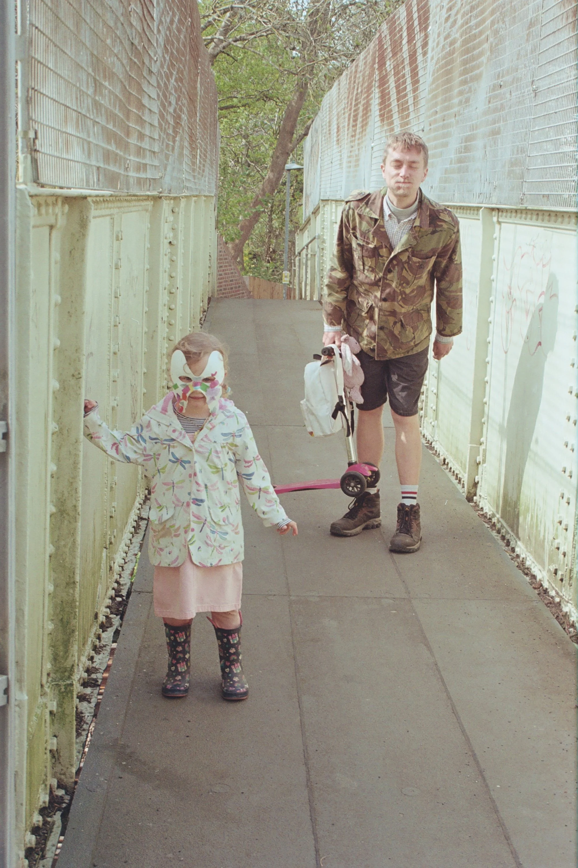 A young girl and an adult man walking on a narrow sidewalk among tall, weathered, and graffiti-covered fences. The girl wears a white mask, colorful jacket, pink skirt, and rain boots, holding the fence. The man wears military camouflage jacket, shor