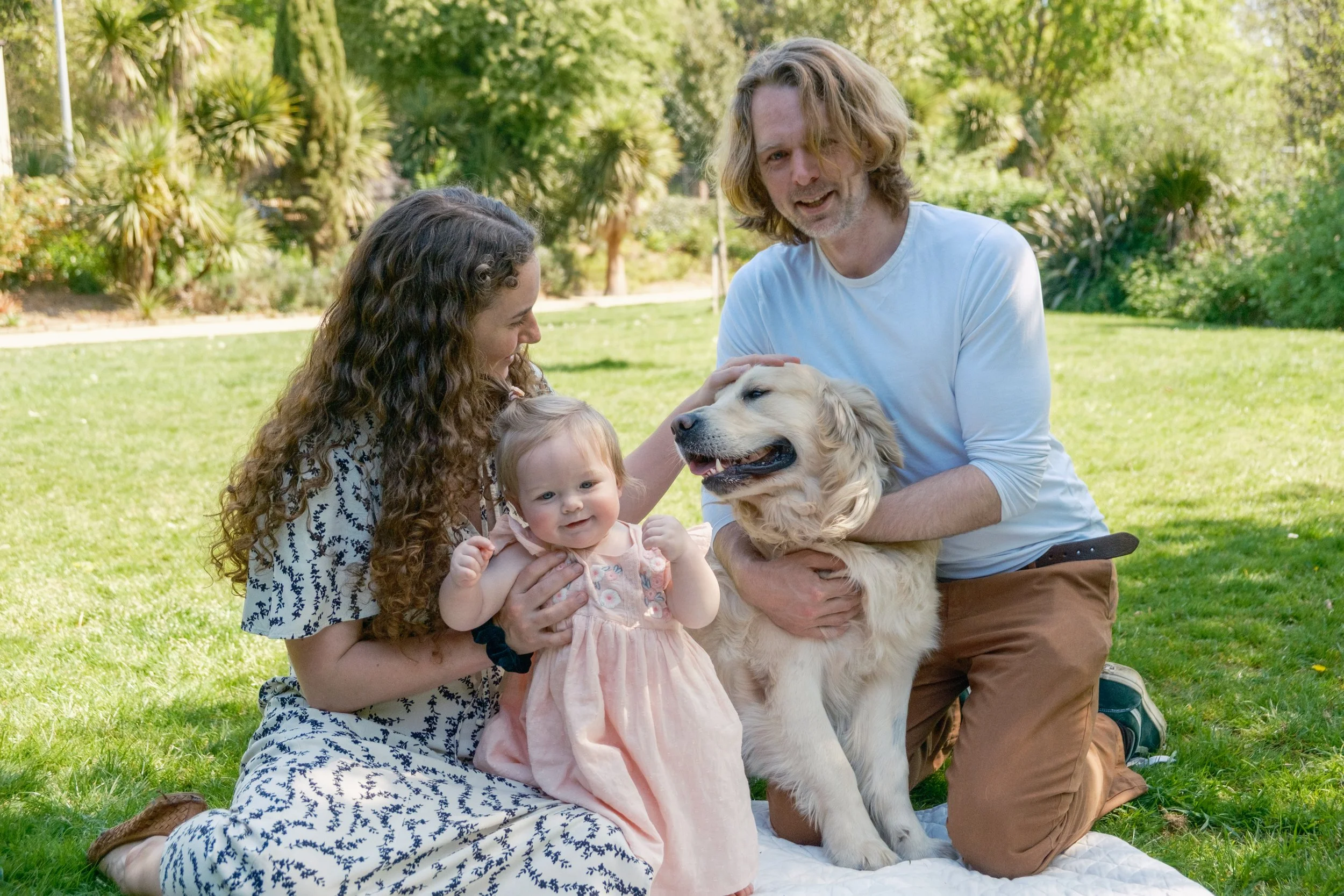 Family of four, including a woman, a man, a young girl, and a puppy, enjoying a sunny day outdoors on grass with trees and bushes in the background.