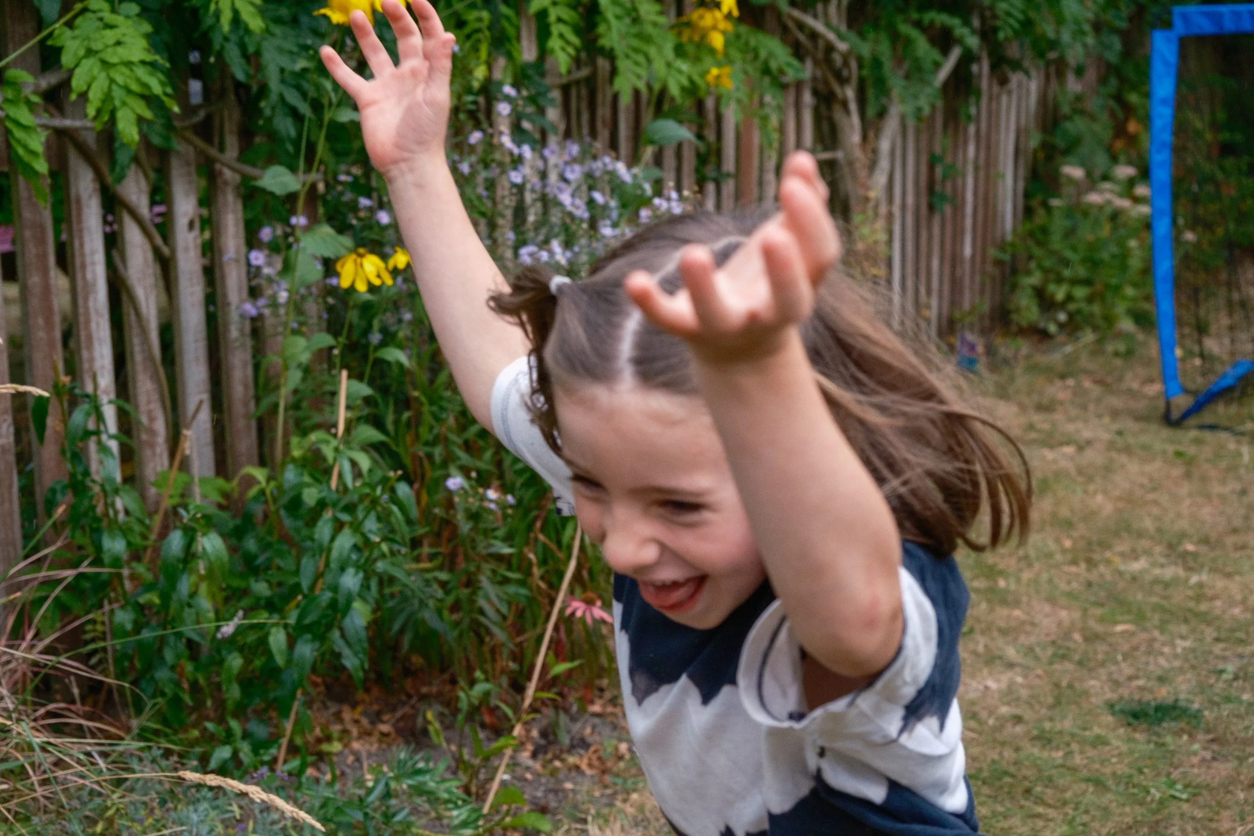 A young girl with brown hair in pigtails is outdoors with arms raised, smiling and playing in a garden with plants, flowers, and a wooden fence in the background.