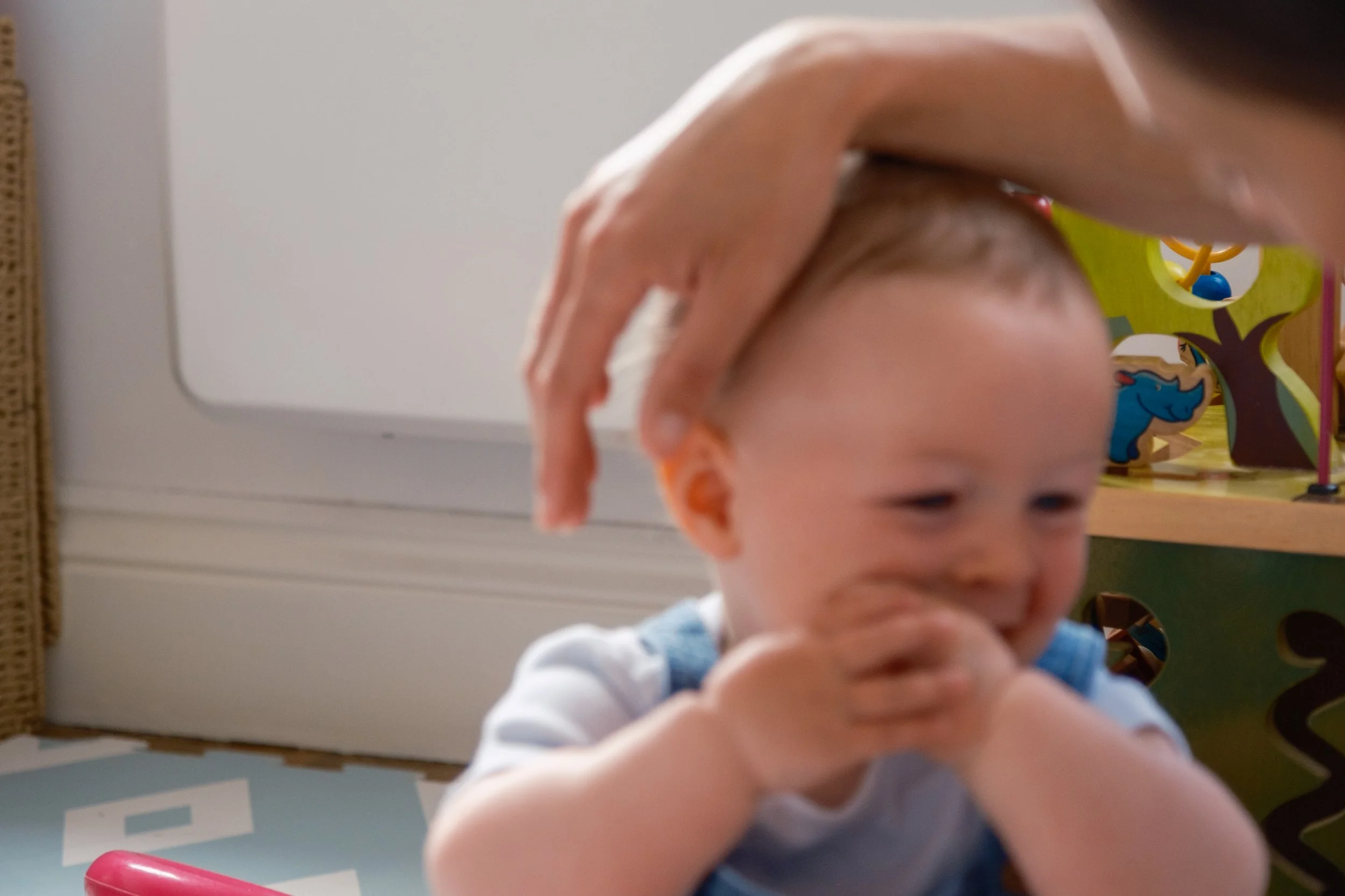 A person gently touching a young boy's head while he is smiling and covering his mouth with his arms, in a room with children's toys in the background.