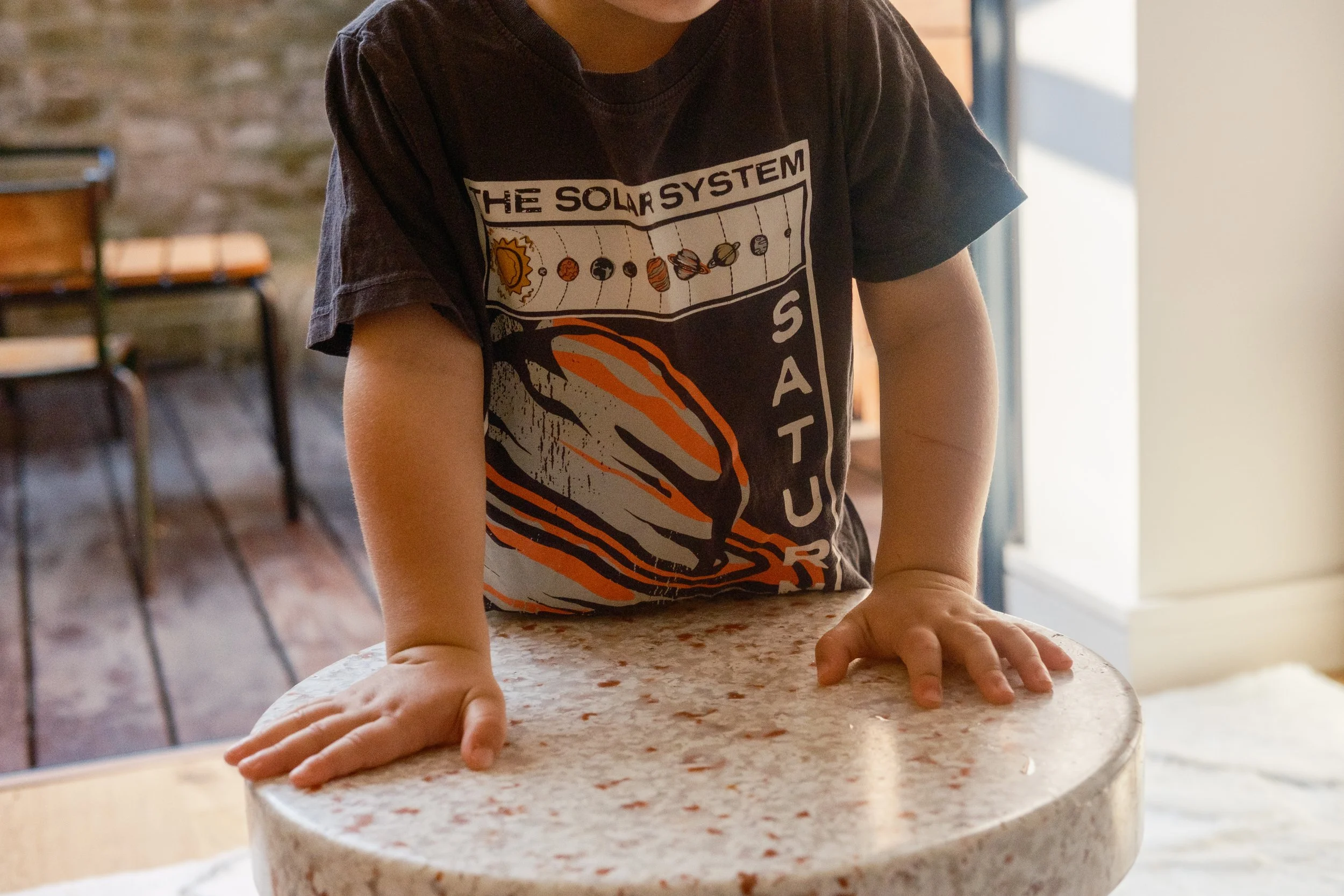 Child leaning on a round kitchen table, wearing a black t-shirt with a solar system graphic and the word 'SATURN' visible.