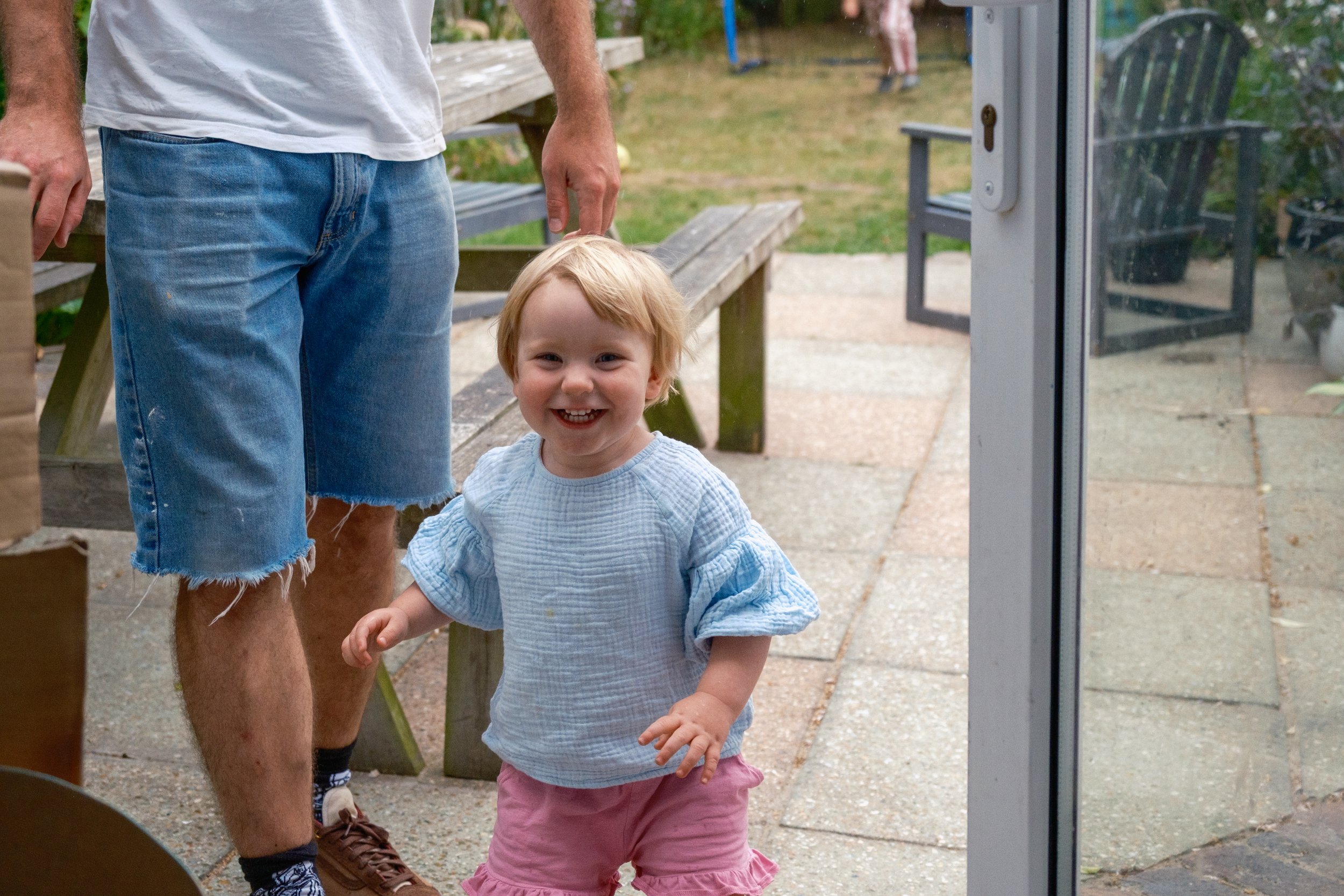 A young girl with short blonde hair smiling and running outside on a stone patio while a man, partially visible, stands nearby. The girl is wearing a light blue top and pink shorts. In the background, there is a wooden picnic table and some greenery.