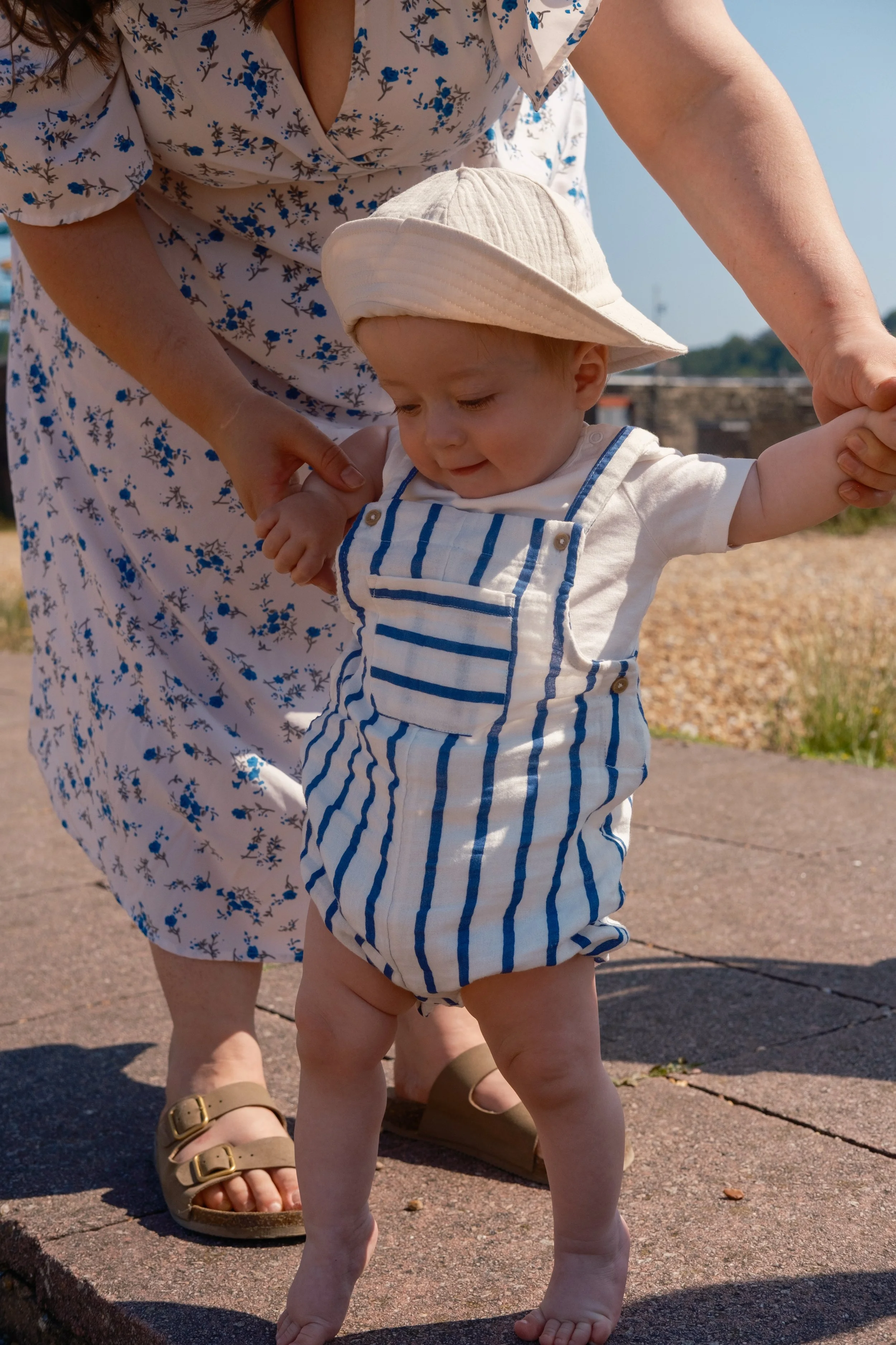 A young child taking steps outside with help from an adult, wearing a sun hat, striped overalls, and a white shirt, on a sunny day.