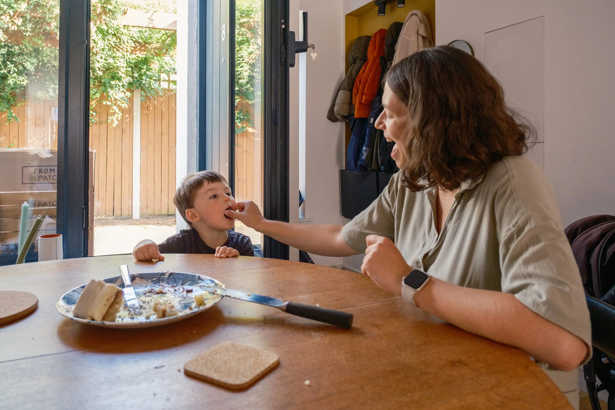 A woman feeding a young boy at a dining table indoors, with the door open to a backyard with a wooden fence and greenery outside.
