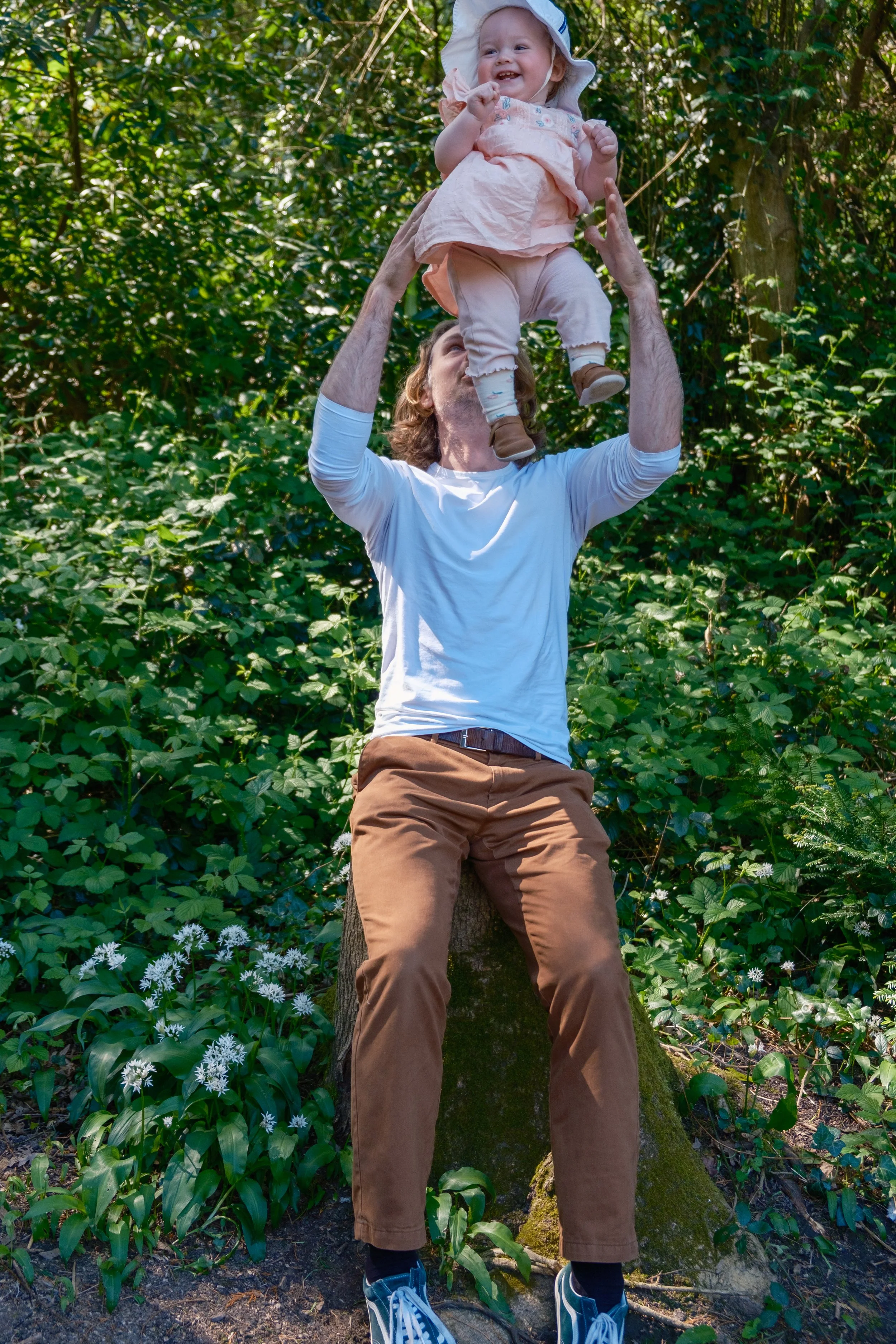 A man sitting on a tree stump in a green forest, holding a smiling toddler girl up in the air.