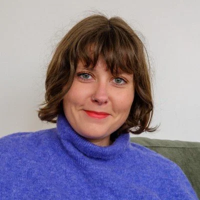 A woman with short brown hair, wearing a purple sweater, smiling while sitting on a green couch in front of a white wall.