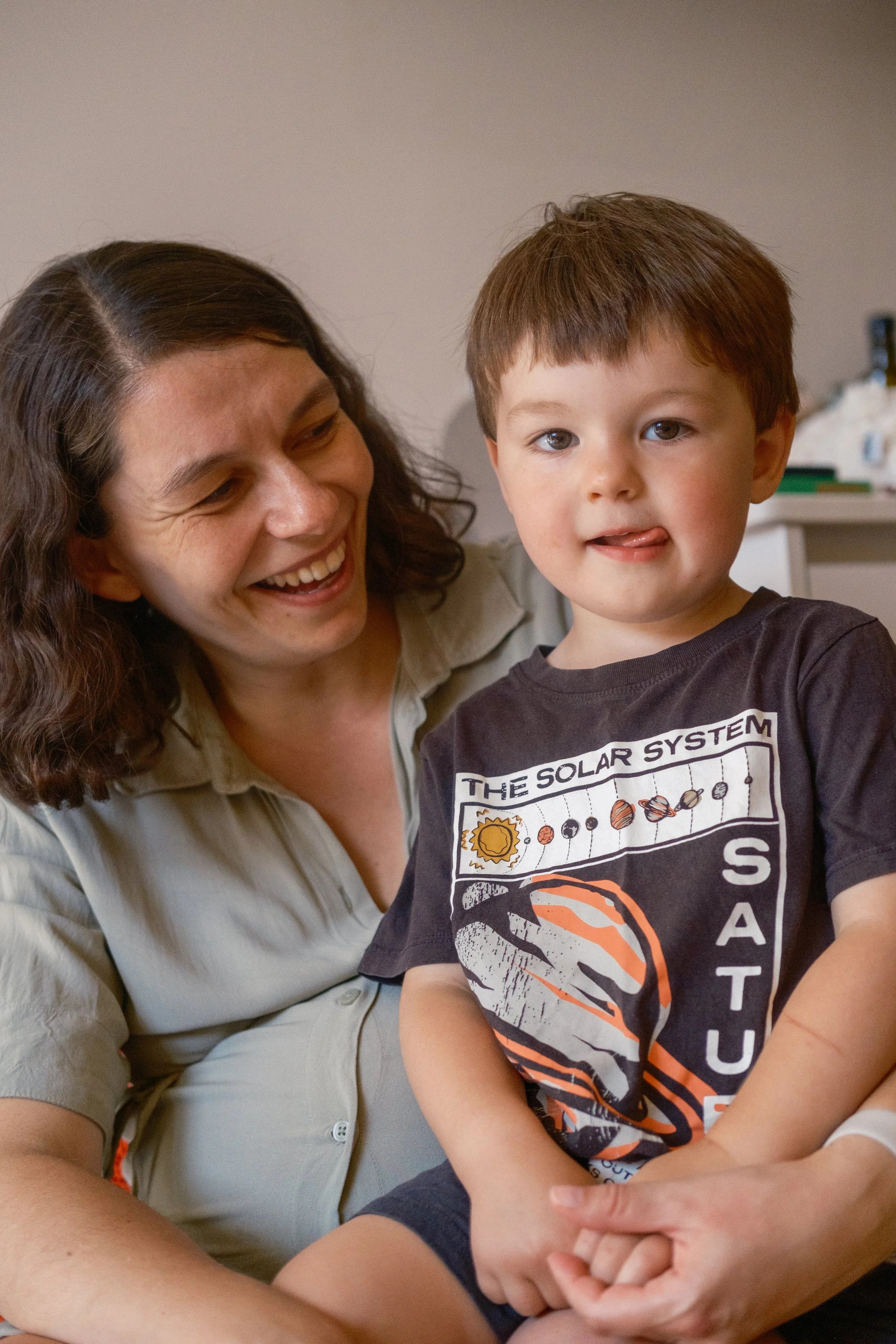 A woman smiling and looking at a young boy, who is sitting on her lap. The boy is sticking out his tongue slightly and looking at the camera. The boy is wearing a black T-shirt with a solar system graphic. The background is a plain wall with some ite