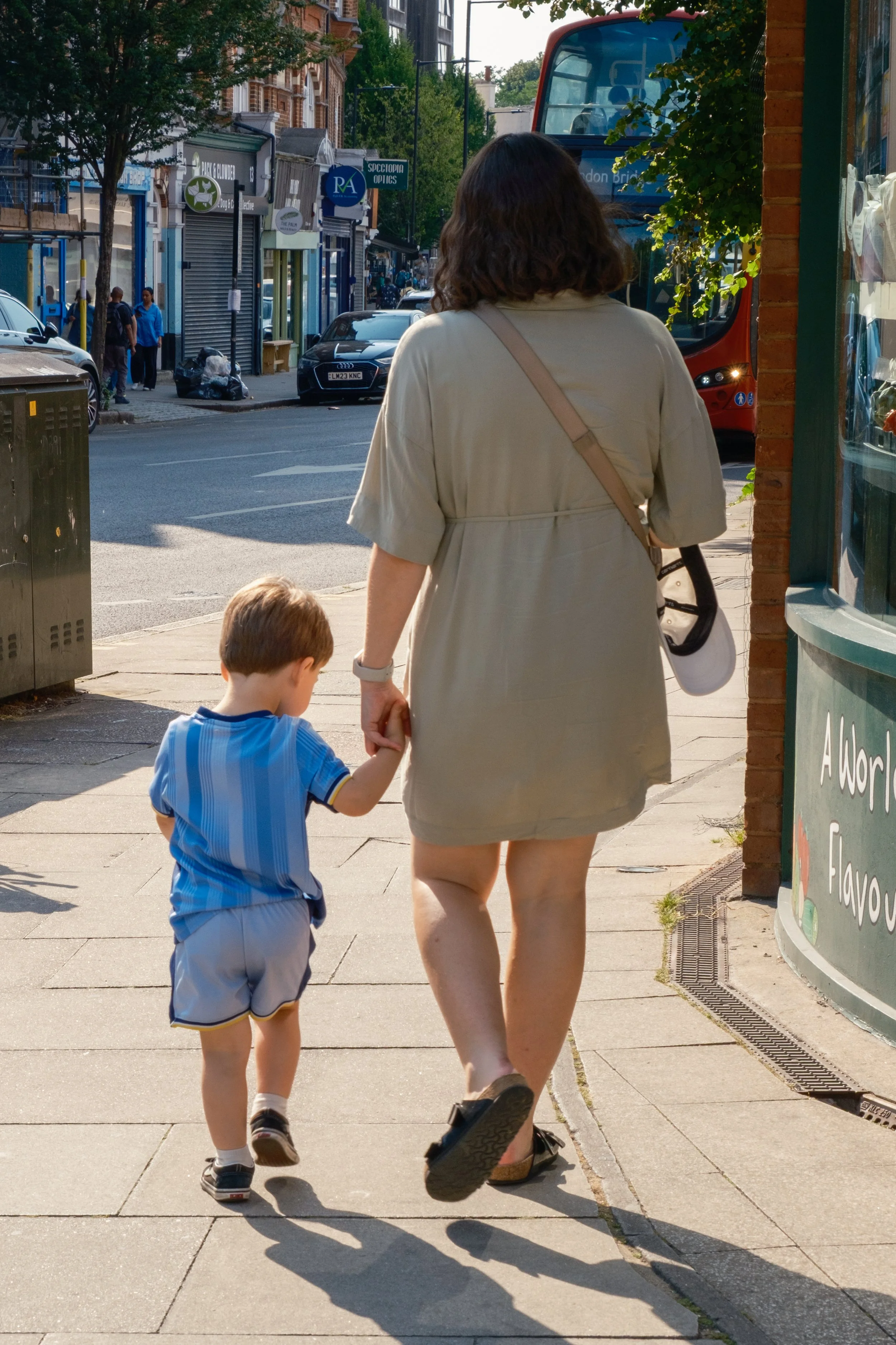 A woman and a young boy walk hand-in-hand on a city sidewalk, with stores and parked cars in the background. The woman is wearing a beige dress and sandals, and the boy is in a blue sports jersey and shorts.