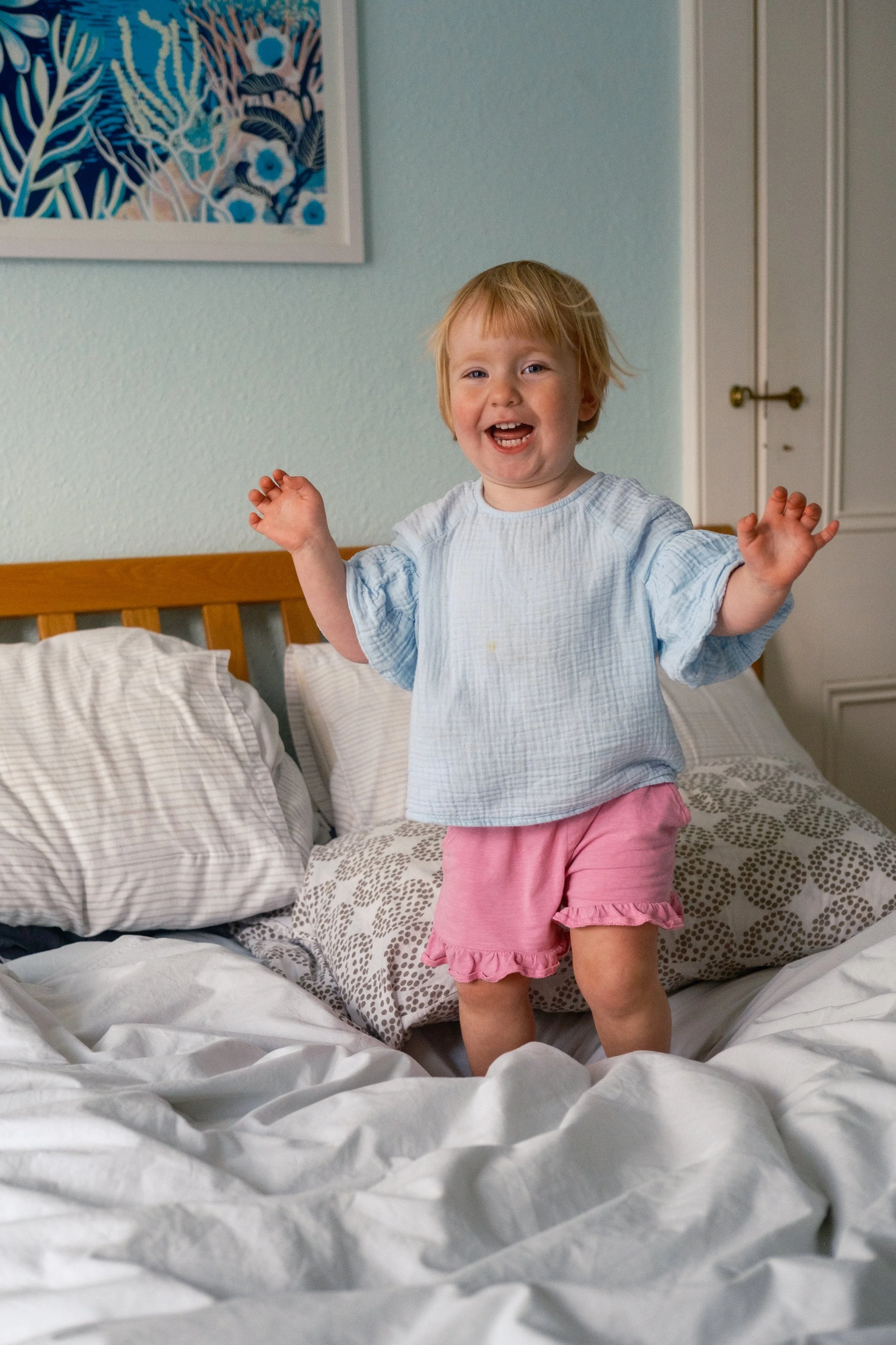 A young girl with blonde hair wearing a light blue top and pink shorts standing on a bed and smiling.