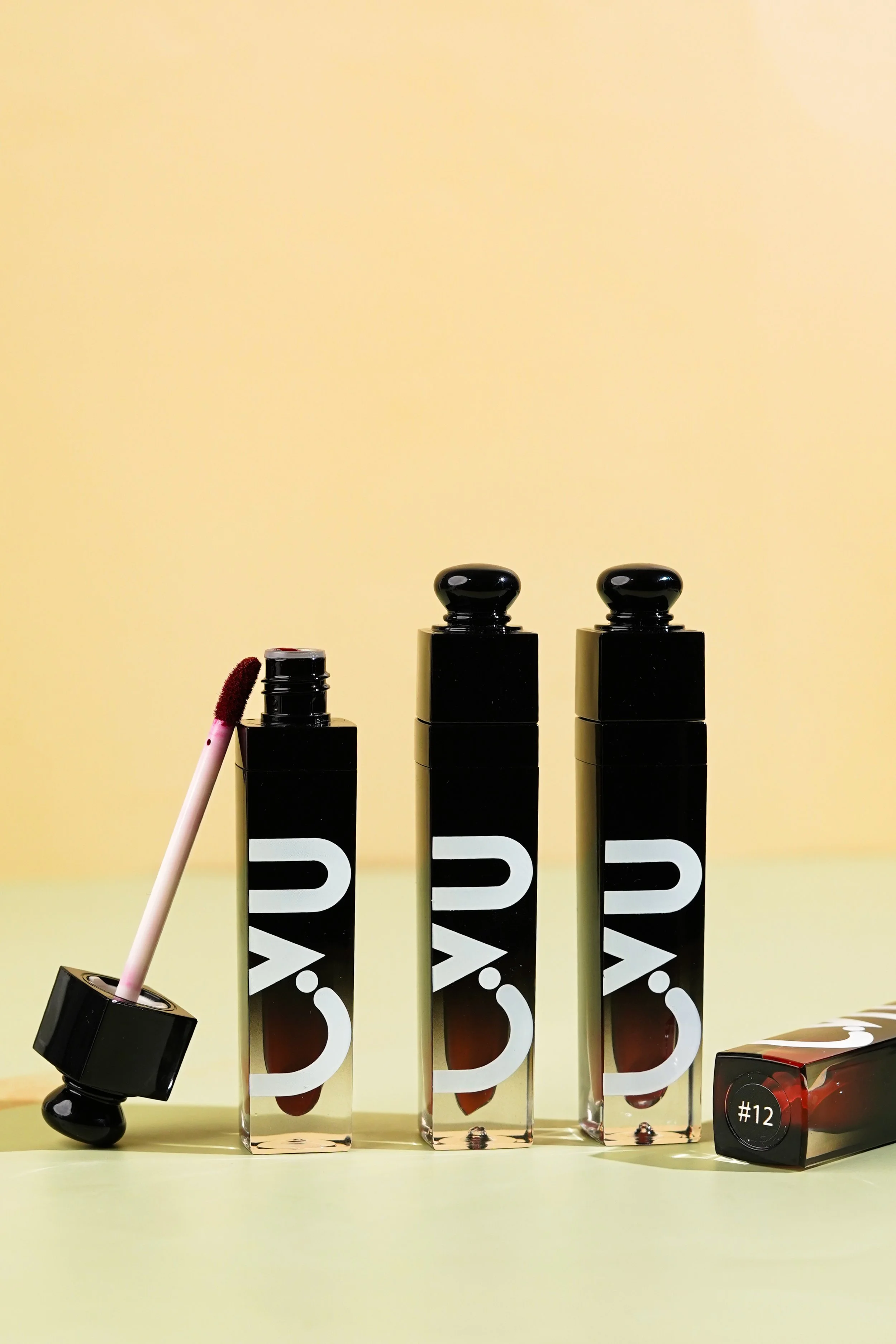 Three black lipstick tubes with white logo, one open with applicator wand, positioned on a light yellow surface against a yellow background, and a closed lipstick tube on the right with a '#12' label on the cap.