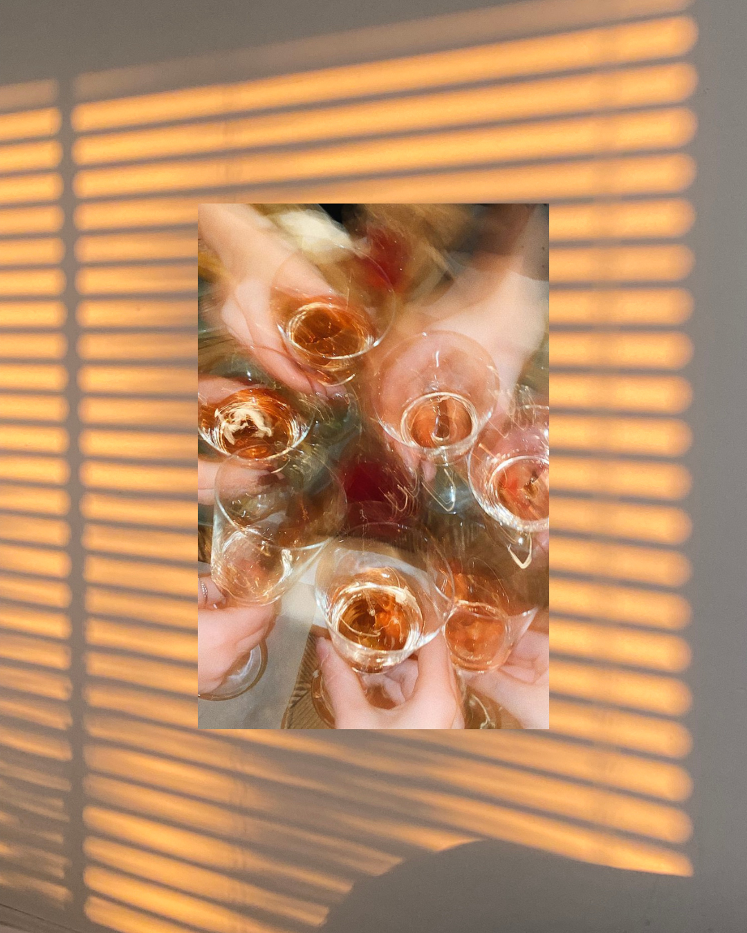 Multiple glasses of a golden-colored beverage being clinked together in a toast, with a shadow pattern from window blinds cast on a surface around the glasses.