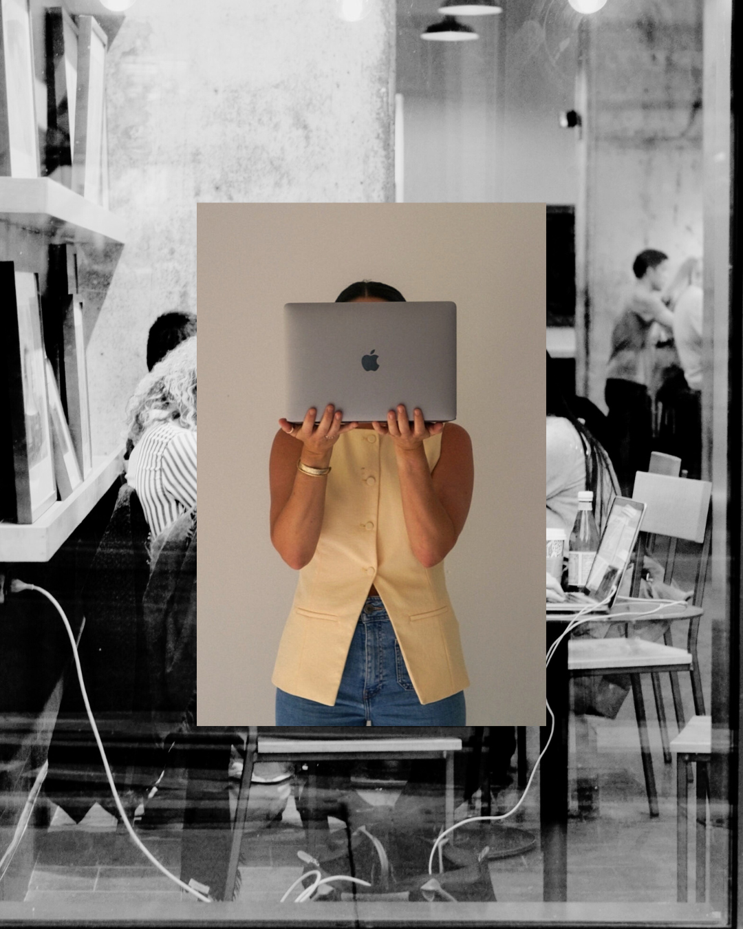 Person taking a selfie with a MacBook in front of their face, wearing a sleeveless yellow vest and blue jeans in a coffee shop or workspace with other people in the background.