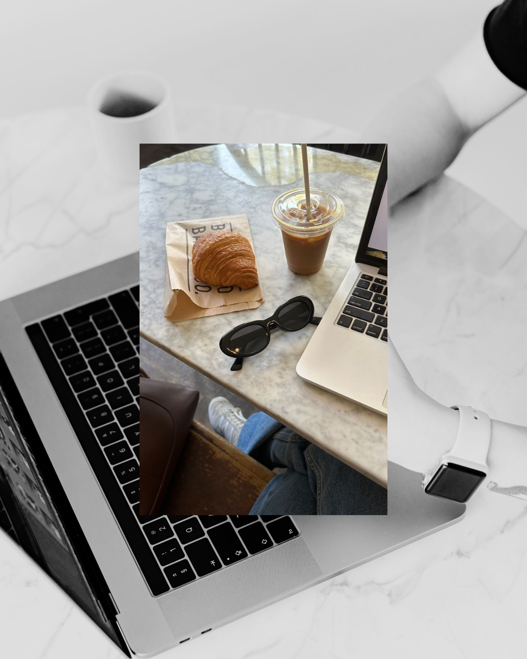 Overlapping images of a coffee cup, a croissant on a paper bag, sunglasses, a laptop, and a person's arm wearing a smartwatch, all on a marble table in a coffee shop.