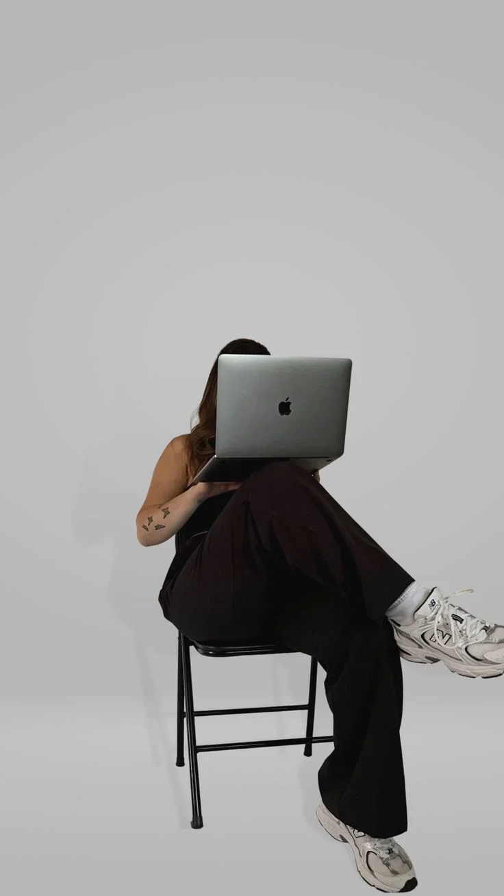 Person sitting on a chair with legs crossed, holding an Apple MacBook in front of their face, against a plain white background.