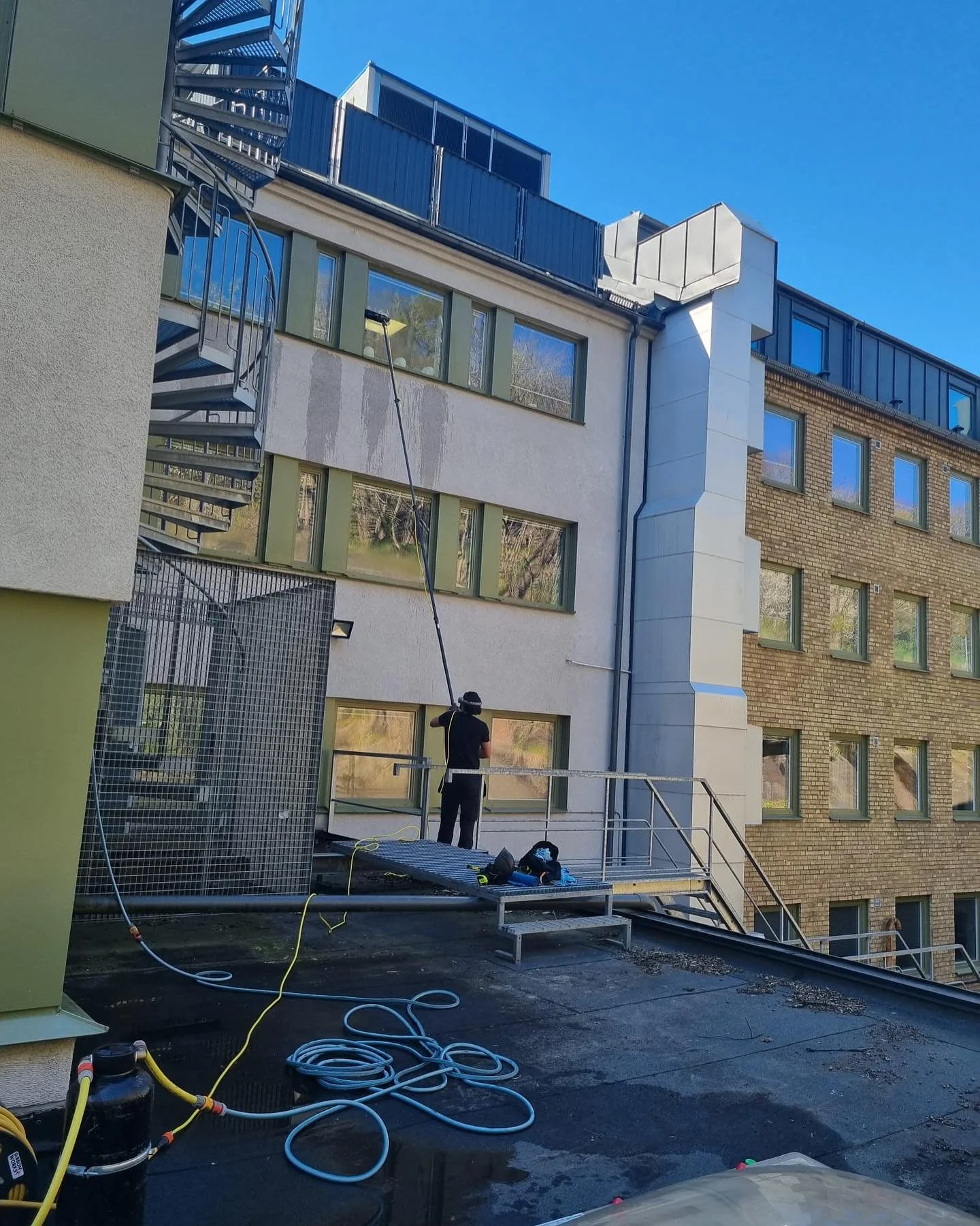 En person som vasker vindu på utsiden av en høy bygning med en lang stang. Det er en dag med klar blå himmel, og det er utstyr som bøtte, skum og lange slanger på taket.