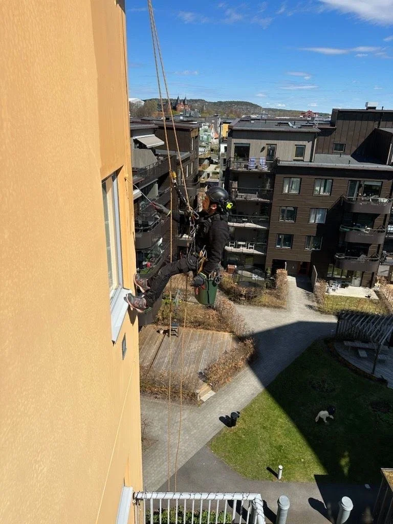 En person i hjelmer og verneutstyr som utfører redningsarbeid fra en boble på siden av en gul bygning i en borettslagsgård.