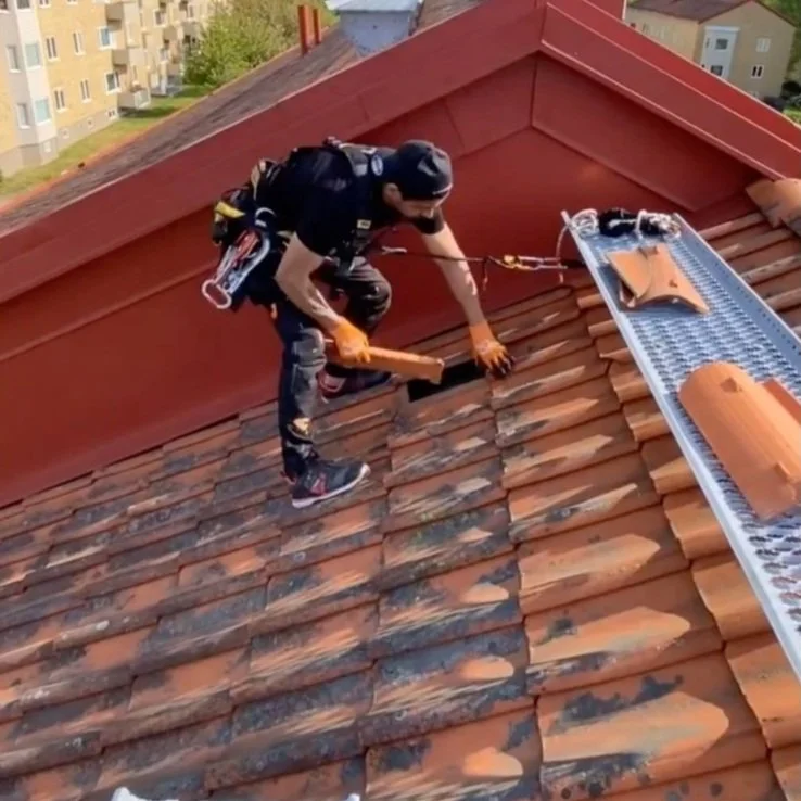 Taktekker som jobber på taket, bruker sikkerhetsutstyr og et brett med takstein, med bygg i bakgrunnen.