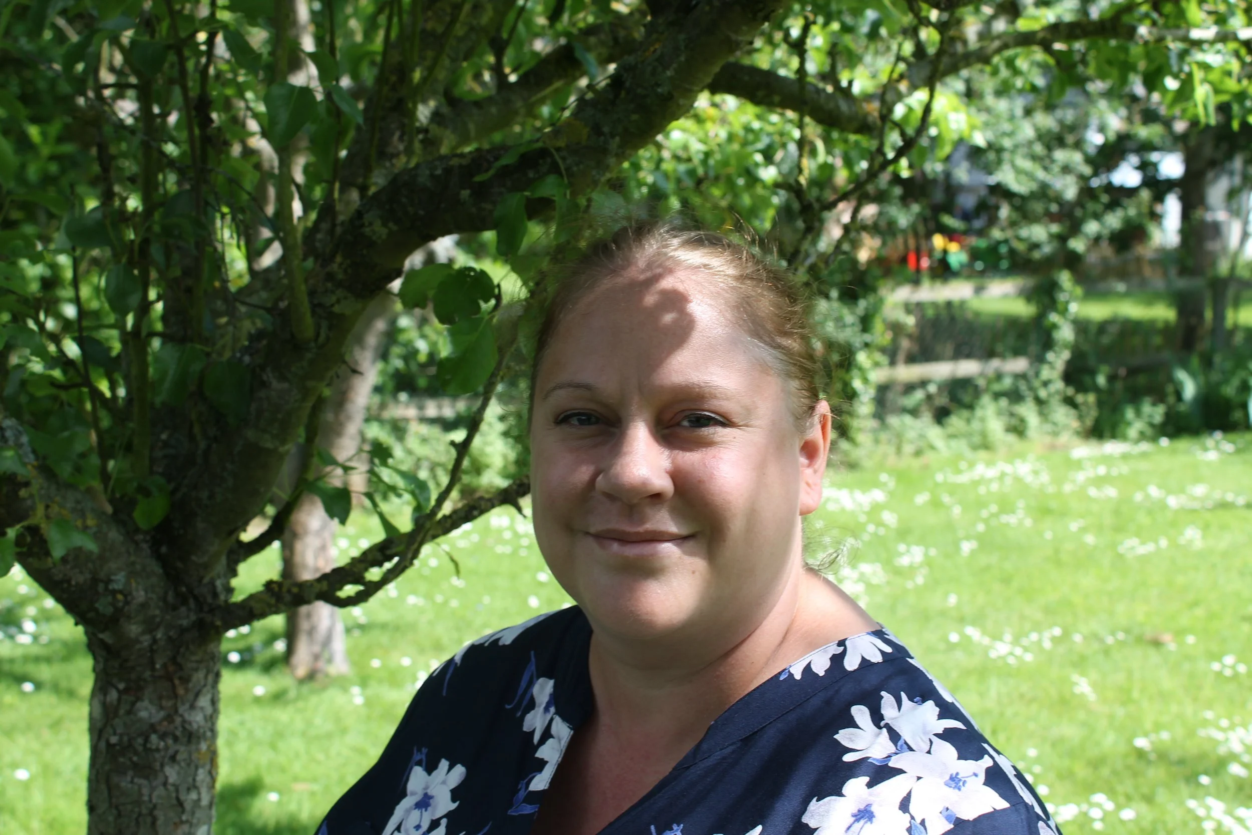 Sarah Bones smiling outdoors beside a tree in a green garden.