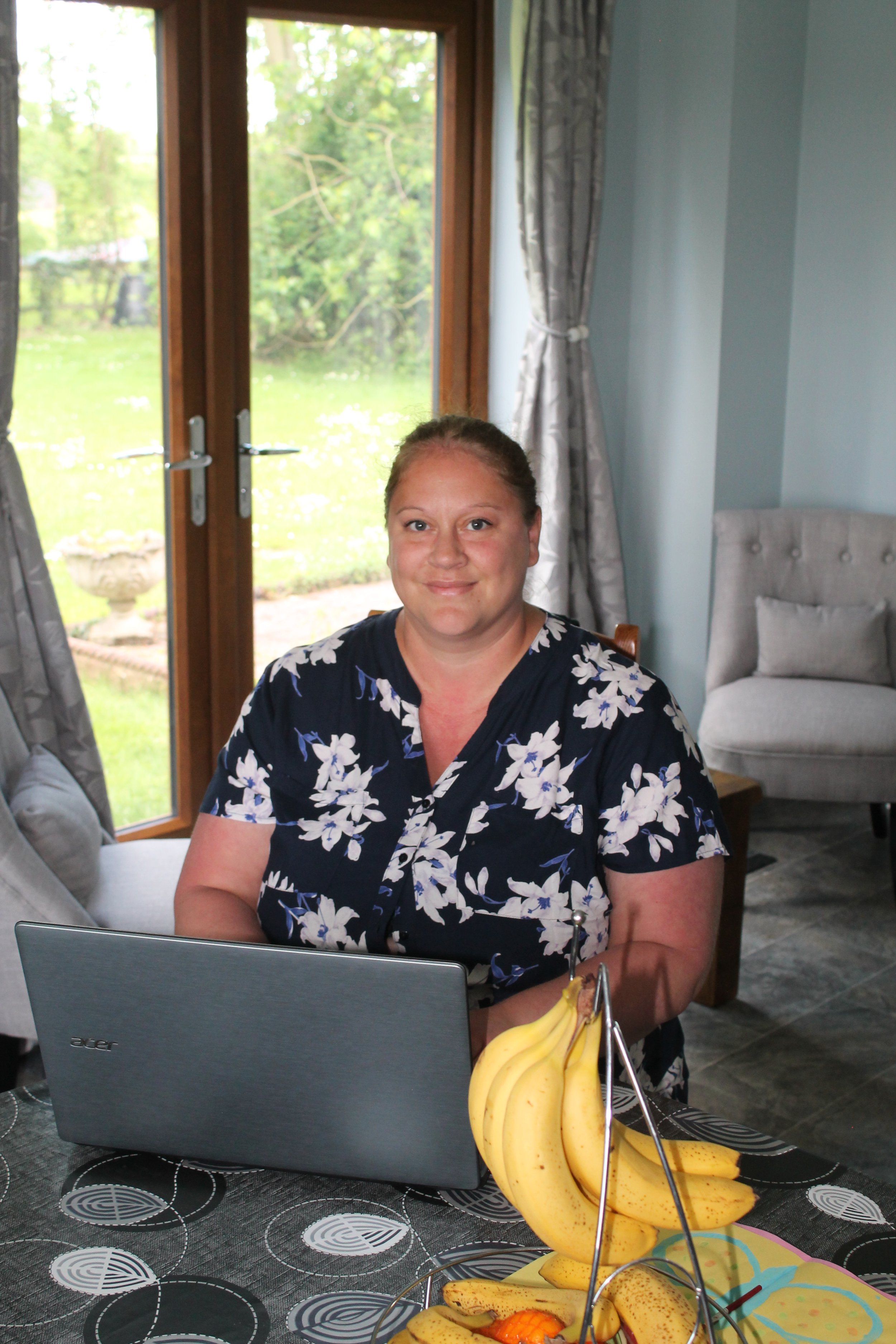 A woman sitting at a dining table with a laptop, bananas, and a fruit platter, inside a room with large glass doors and a view of a green backyard.