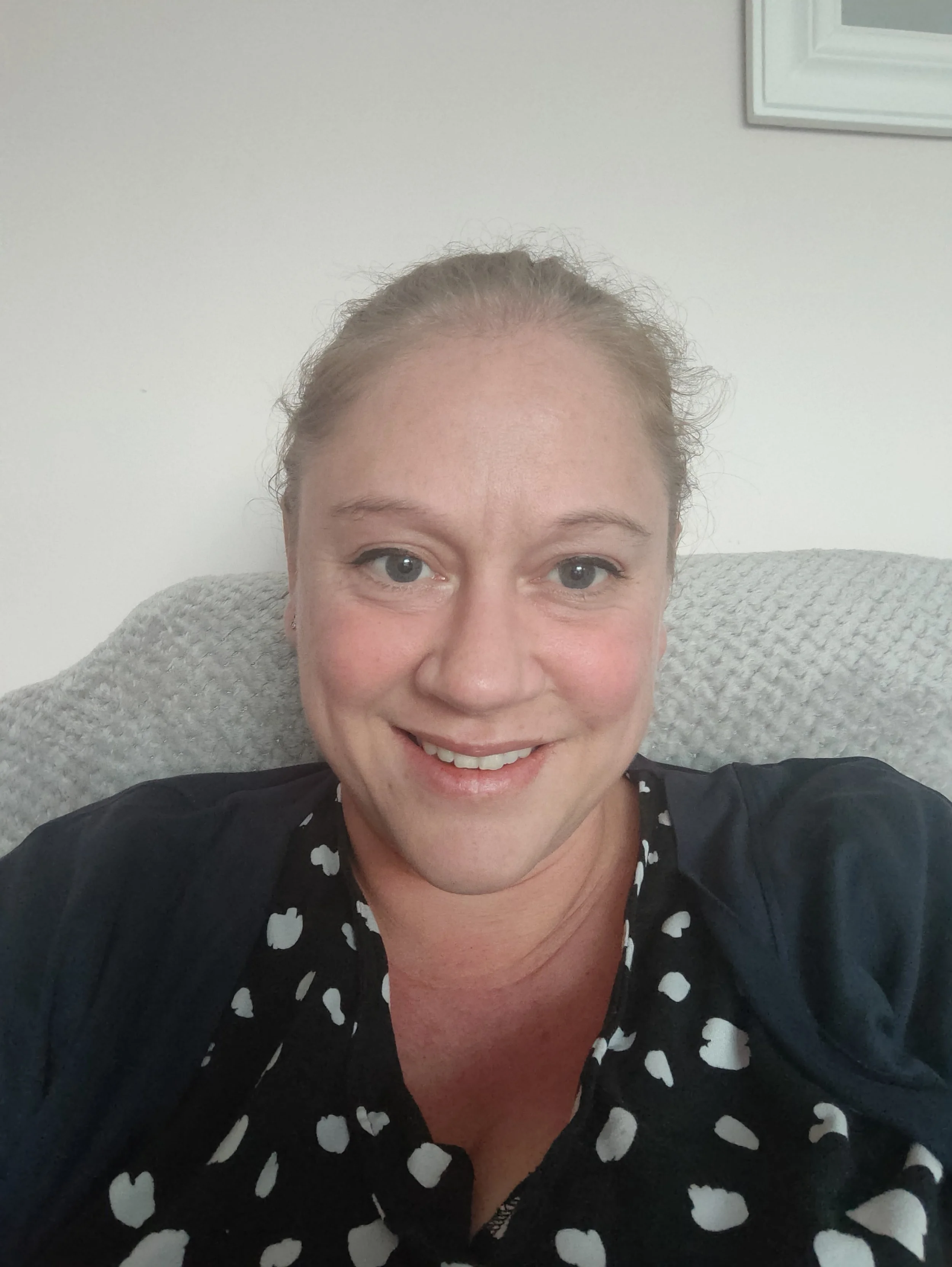 Sarah Bones smiling in a close-up selfie, sitting on a grey upholstered chair with a white wall and a partial picture frame in the background.
