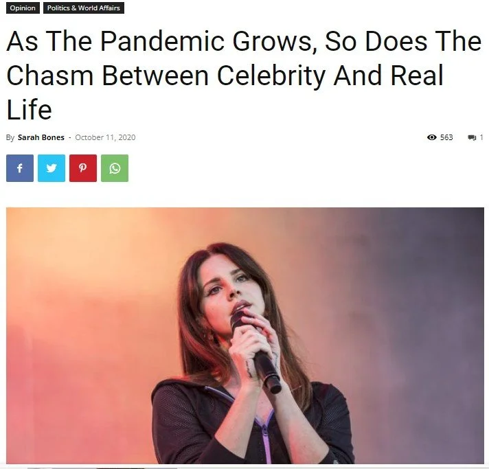 A woman with long dark hair, wearing a black jacket, holds a microphone close to her mouth, appearing to speak or sing on stage against a colorful background.