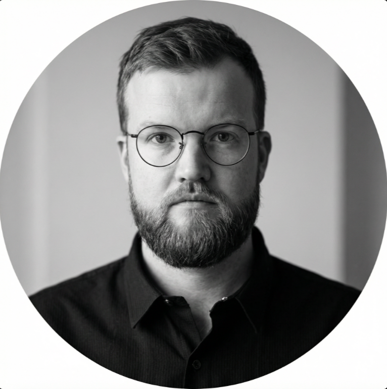 Black and white portrait of a man with glasses, a beard, and short hair, wearing a dark shirt.