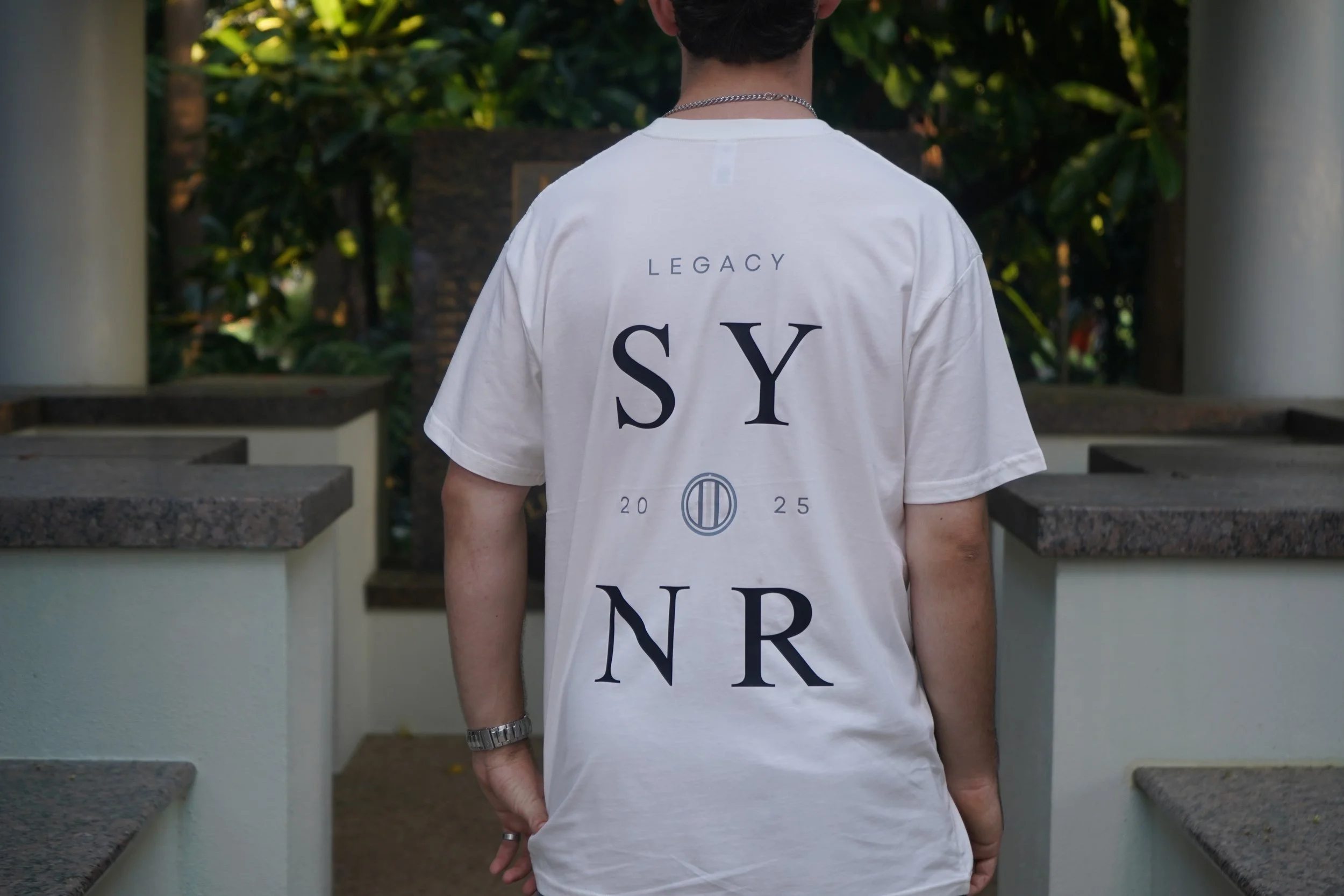 A person with short dark hair wears a white T-shirt with the words 'LEGACY SYN R 2025' printed on the back, standing outdoors between two white columns with greenery in the background.