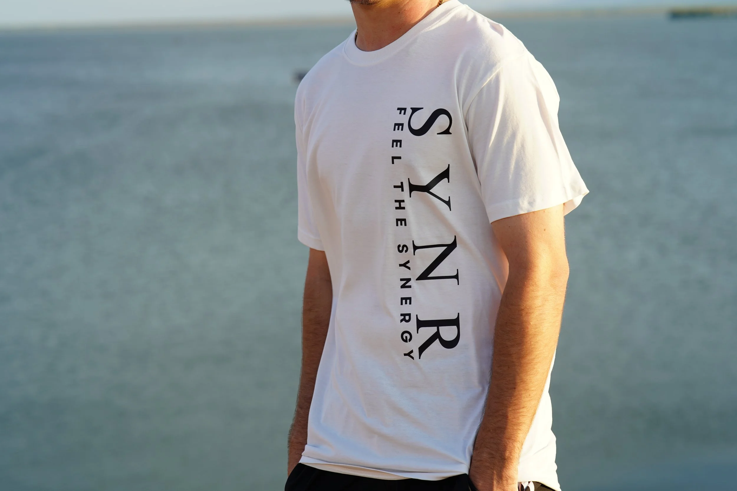 Person wearing a white T-shirt with the words "SYNR" and "FEEL THE SYNERGY" printed on it, standing outdoors near water.