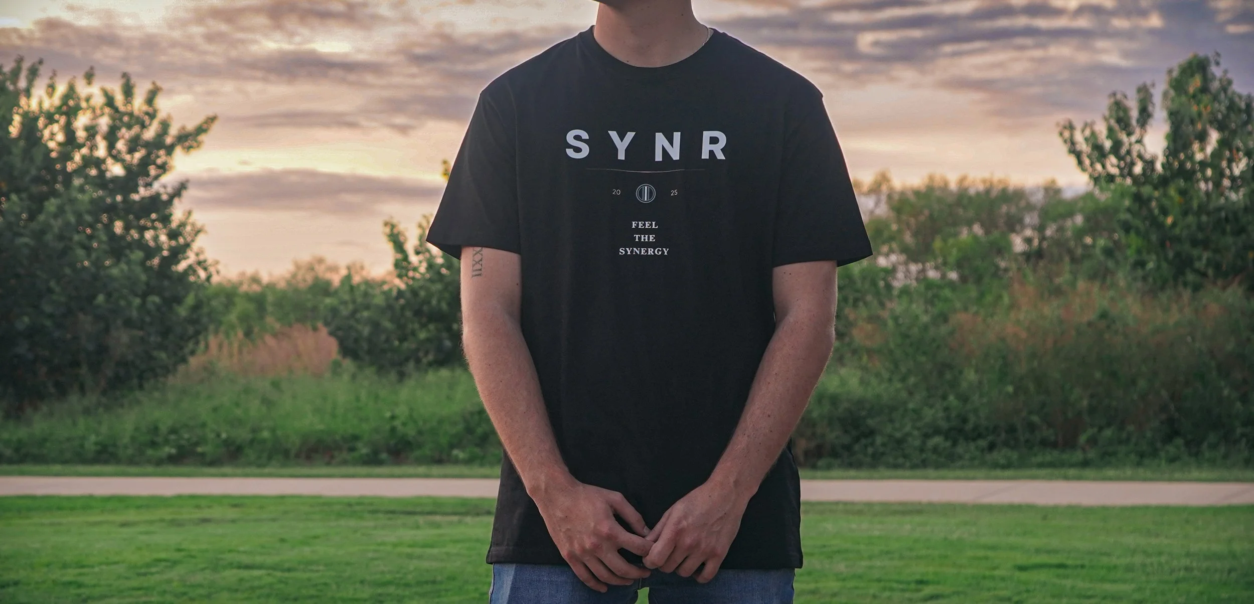 A person standing outdoors at sunset, wearing a black t-shirt with the word 'SYNR' and the phrase 'FEEL THE SYNERGY' printed on it, with trees and grass in the background.