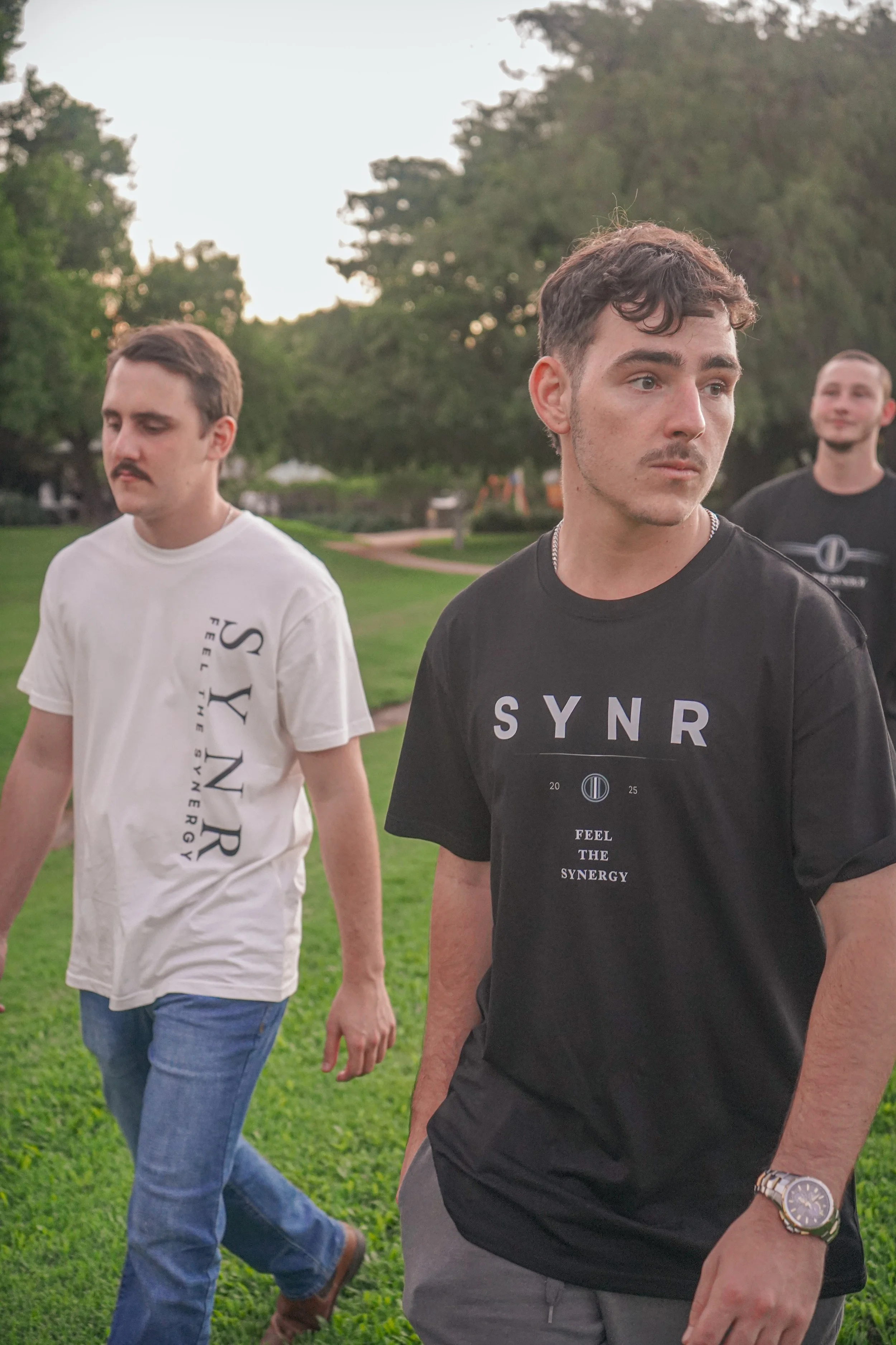 Three young men walking outdoors on a grassy area with trees in the background during sunset. They are dressed casually, with two wearing T-shirts with the word 'SYN' and 'SYNR' printed on them.
