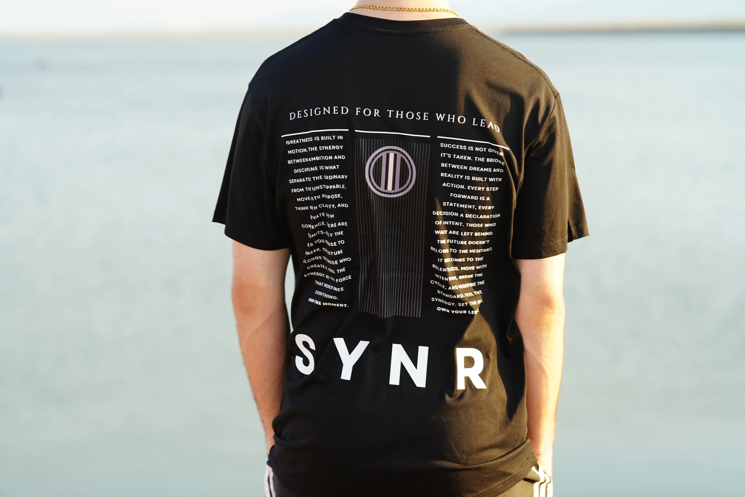 Person wearing a black T-shirt with white text and graphics, standing near a body of water. The T-shirt features the words 'DESIGNED FOR THOSE WHO LEAD' at the top, a circular symbol in the center, and the word 'SYN' at the bottom.