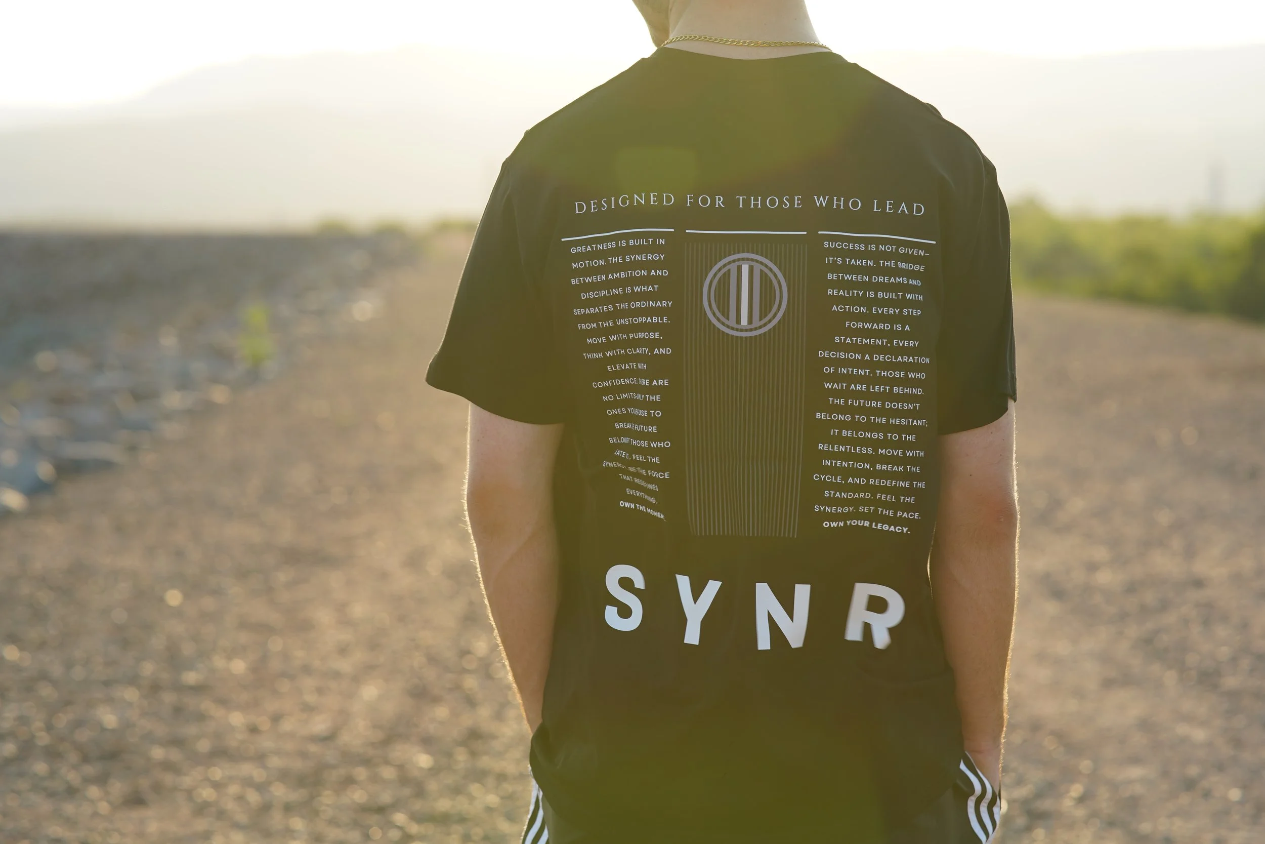 Person wearing a black T-shirt with white text, standing outdoors on a dirt road during sunset, with a mountainous landscape in the background.