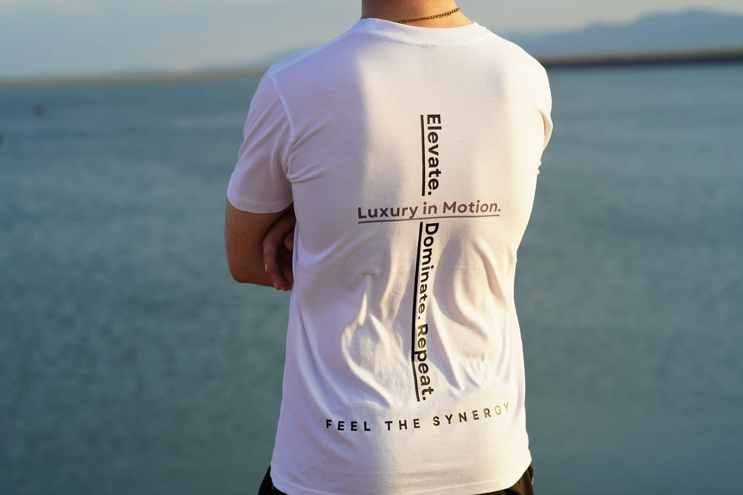 A person wearing a white t-shirt with black and brown text, standing by a body of water during the daytime. The t-shirt has the words "Elevate," "Dominate," "Repeat," and "Feel the Synergy" printed on it.