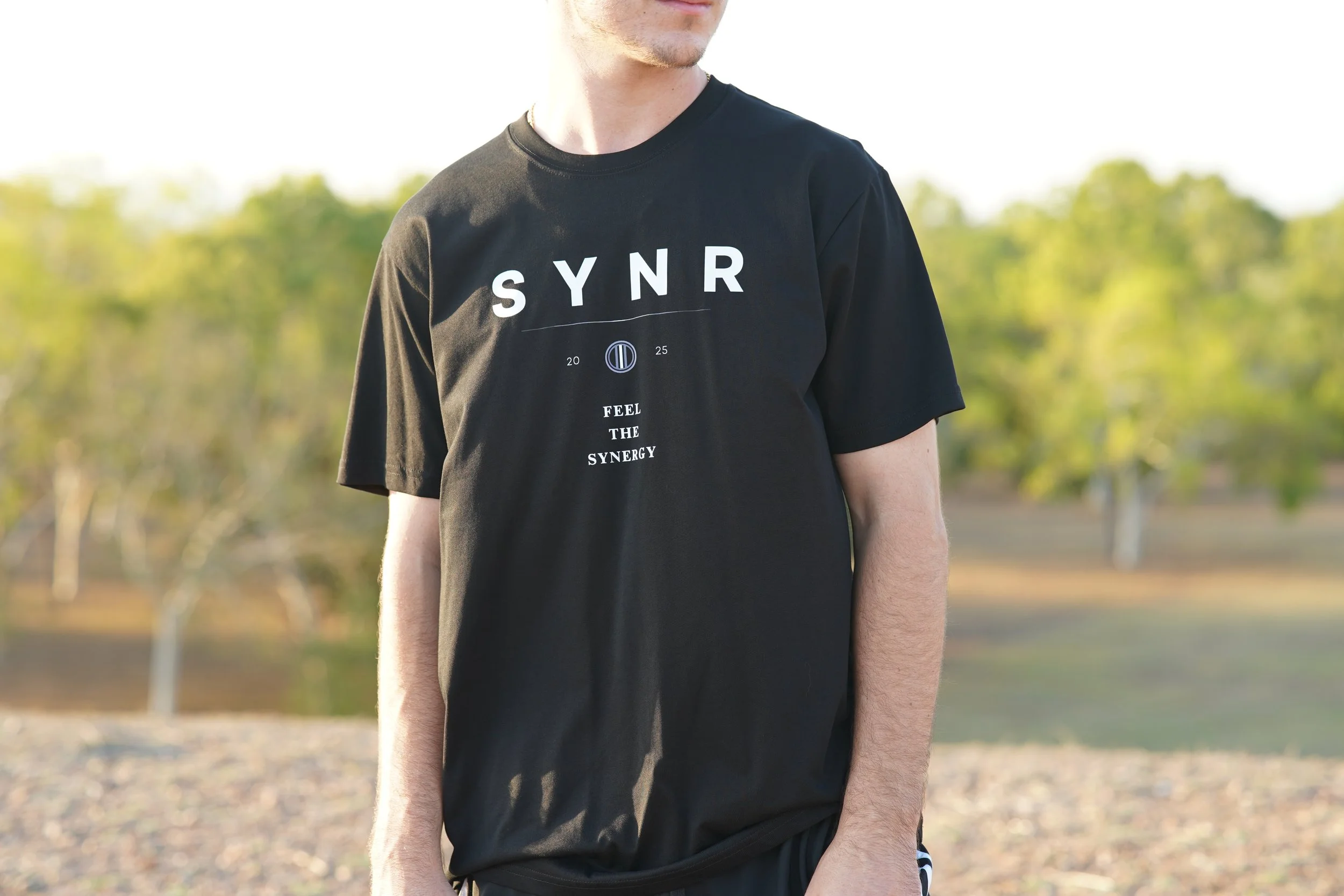 A person wears a black T-shirt with the words 'SYN' and 'FEEL THE SYNERGY' printed on it, standing outdoors in a park or natural setting.