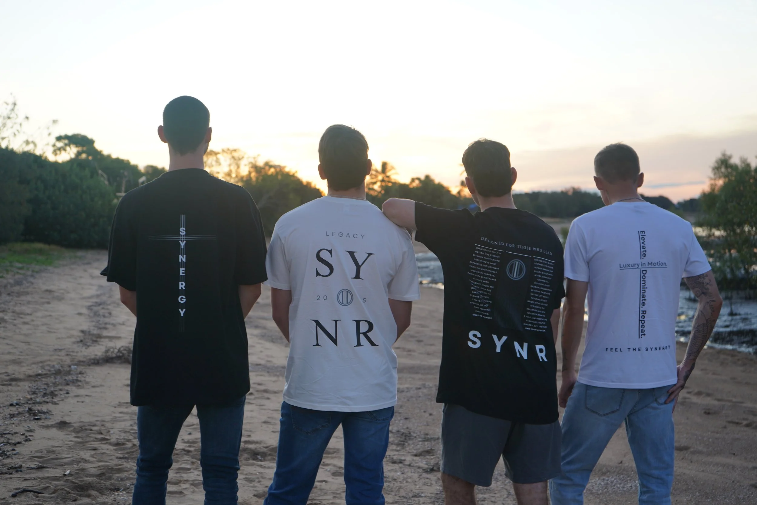 Four young men standing side by side on a beach at sunset, with trees and water in the background, facing away from the camera.