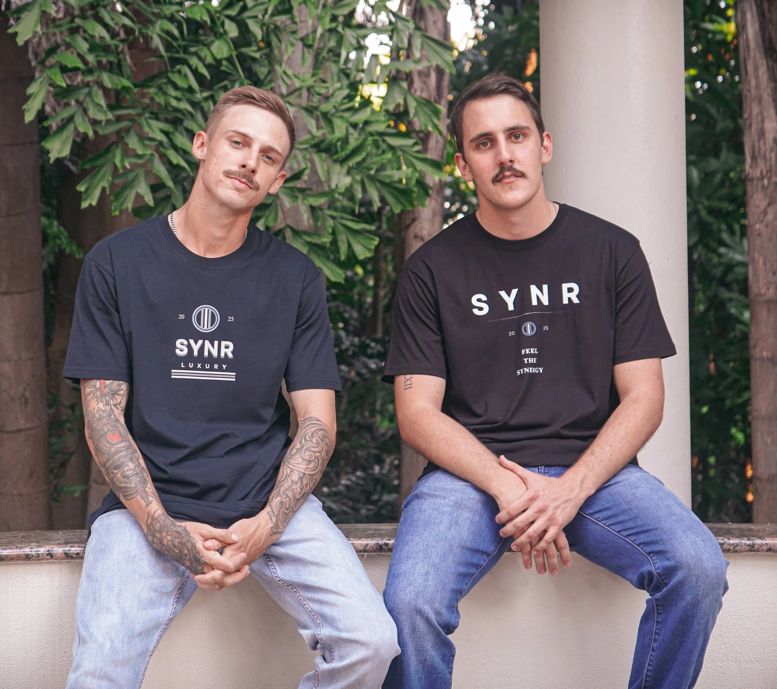 Two men sitting outdoors on a ledge, wearing black "SYNR" t-shirts, with green foliage and trees in the background.