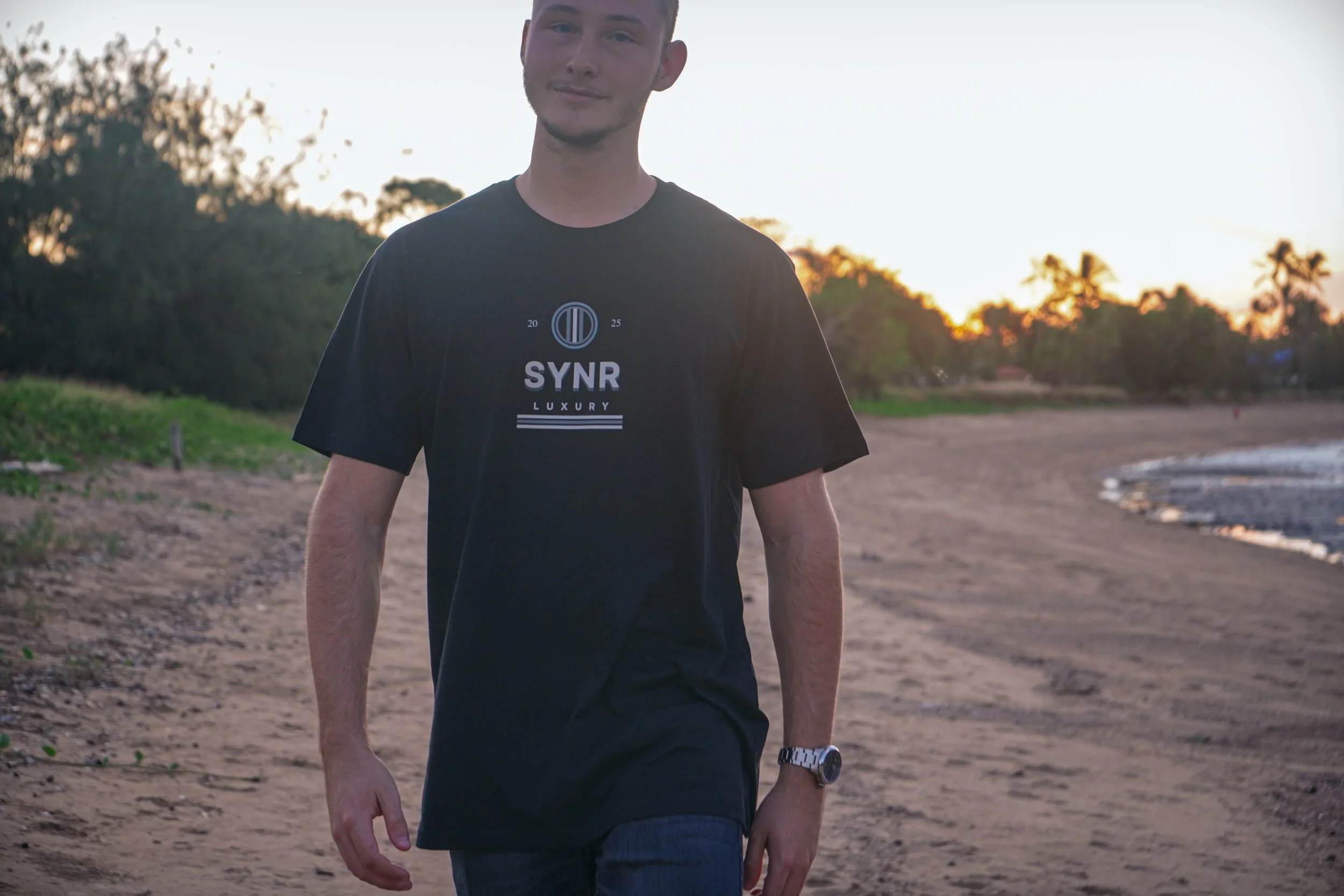 Young man walking on a beach at sunset, wearing a black t-shirt with the word 'SYN' printed on it.